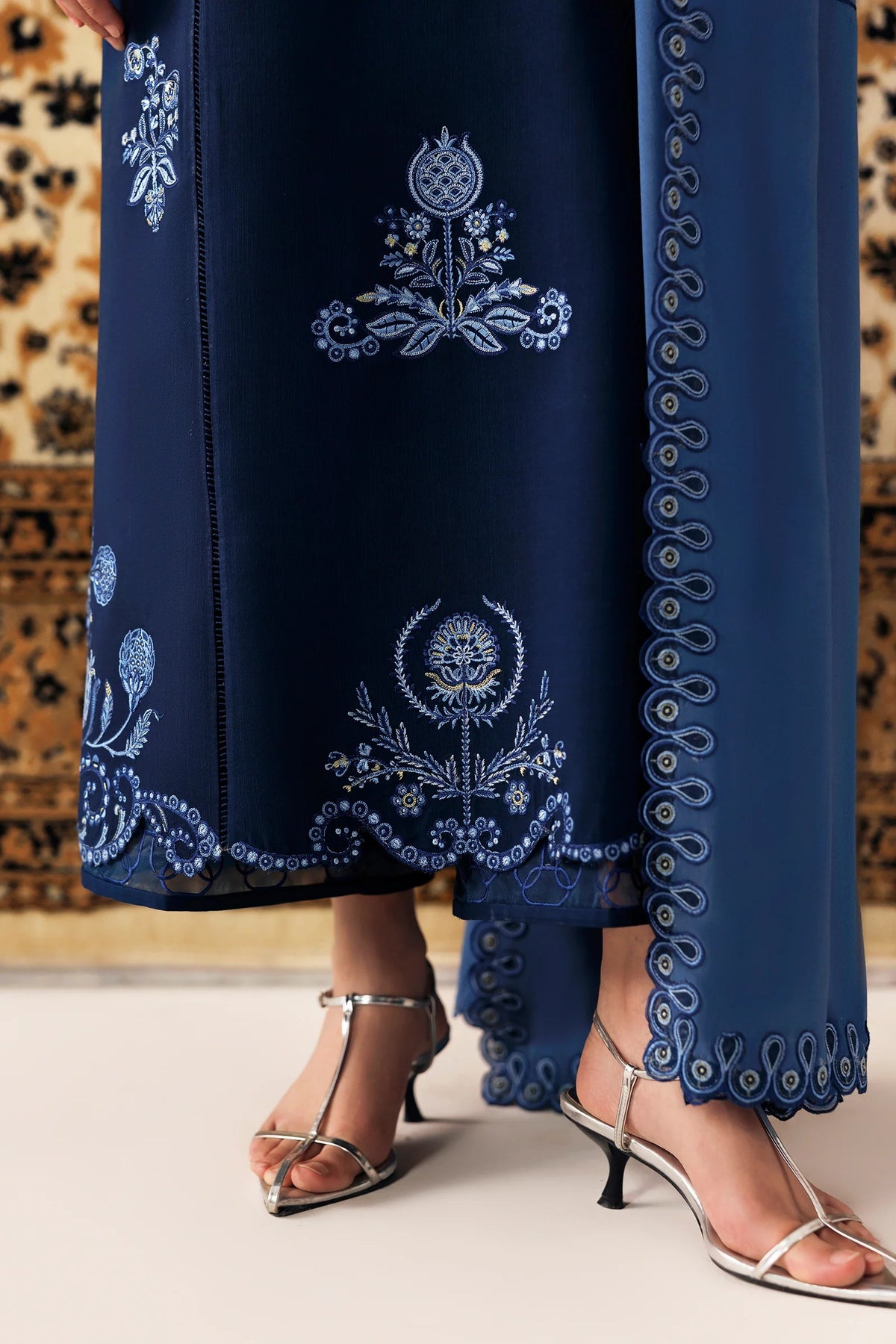 A close-up of a navy blue dress or caftan with intricate light blue and white embroidery, shown on a model wearing silver heeled sandals.