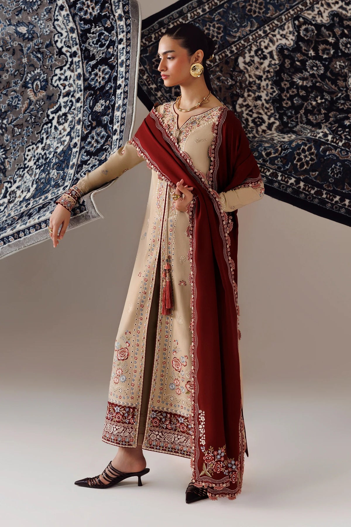 A woman models a traditional South Asian outfit consisting of a beige embroidered kurta, a deep maroon dupatta, and black strappy heels, posed against a studio backdrop with patterned rugs.