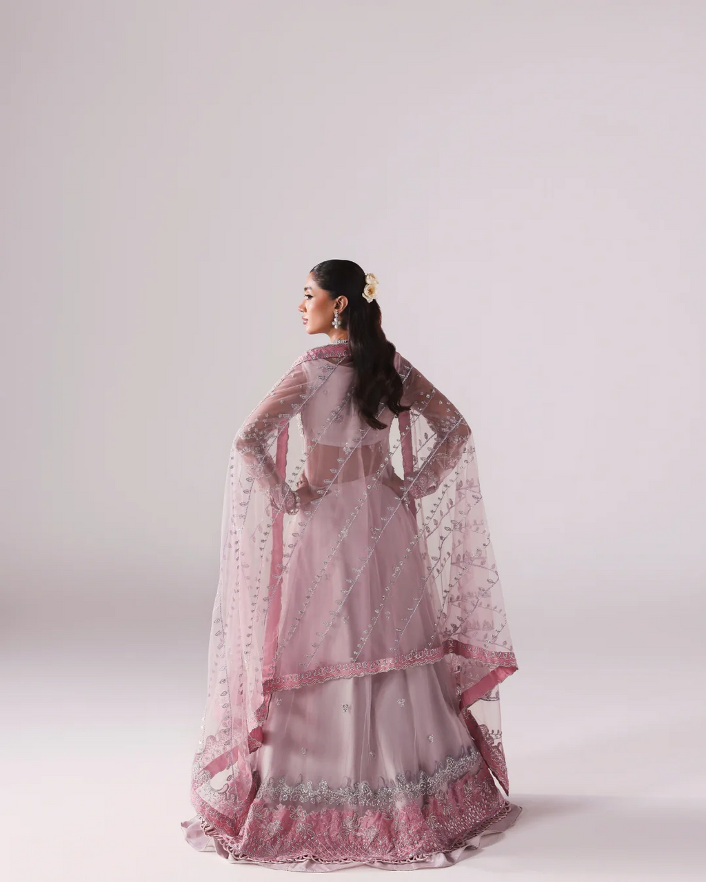 A woman wearing a light pink traditional outfit with a matching sheer embroidered dupatta, photographed from the back against a plain light gray studio background.