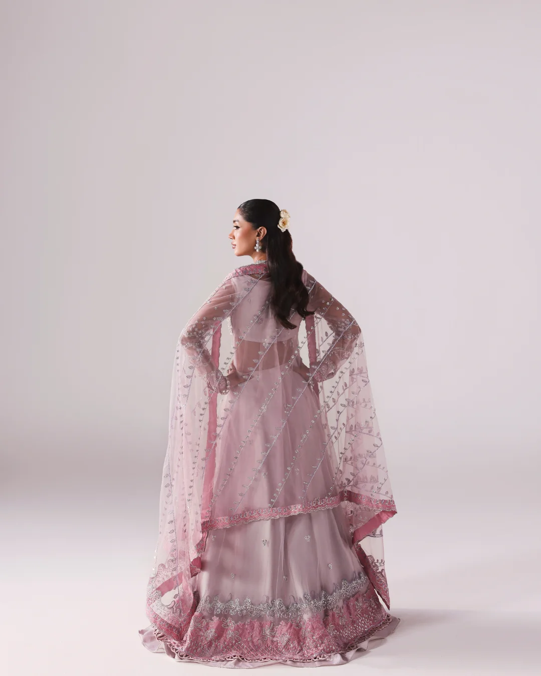 A woman wearing a light pink traditional outfit with a matching sheer embroidered dupatta, photographed from the back against a plain light gray studio background.