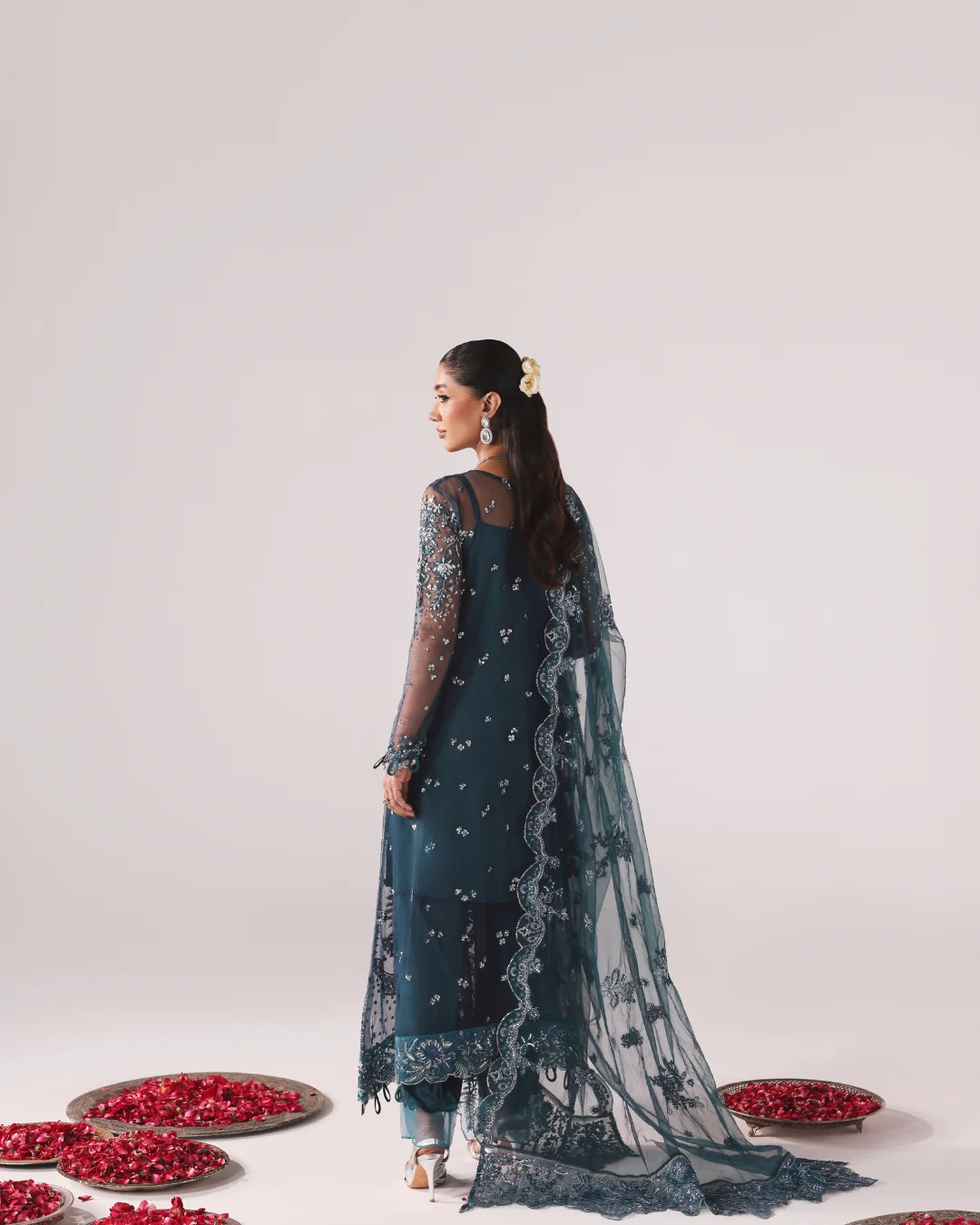 A woman stands in profile wearing a dark teal embroidered traditional outfit with sheer sleeves and a matching embroidered dupatta. The scene is photographed in a clean studio with a light gray background, and several trays of red flower petals are arranged on the floor around her.