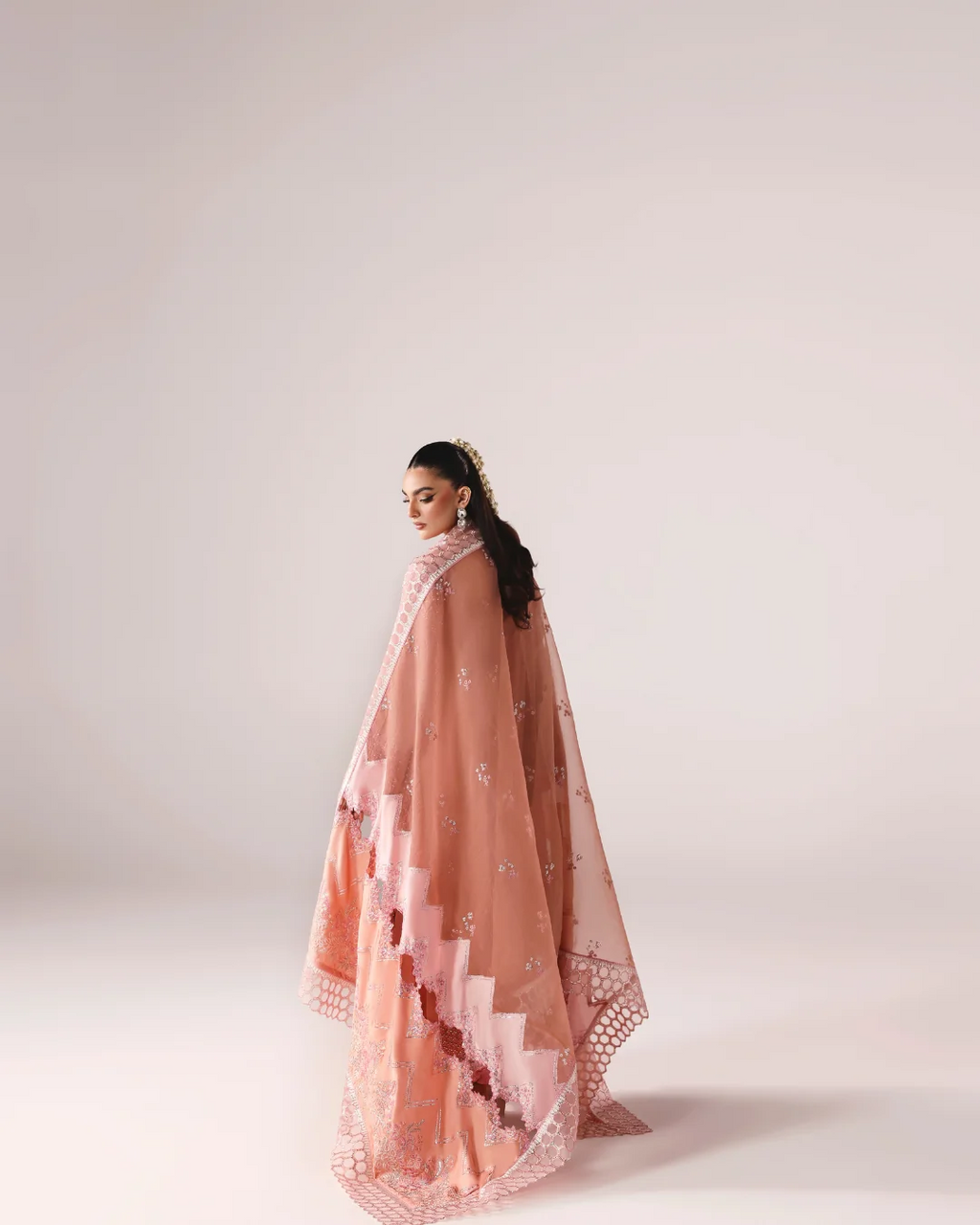 A woman wearing a peach-pink embroidered shawl or wrap with scalloped edges, photographed in profile against a soft light beige studio background.
