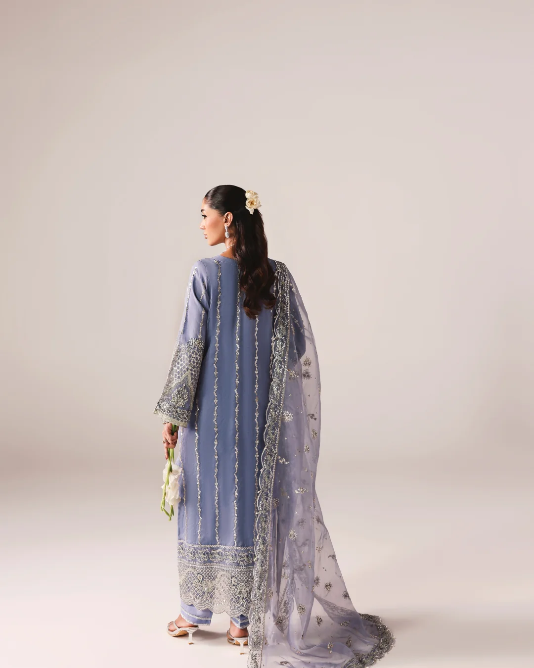 A woman is shown from the back wearing a light blue traditional outfit with silver embroidery and a matching sheer dupatta, set against a plain light beige studio background.