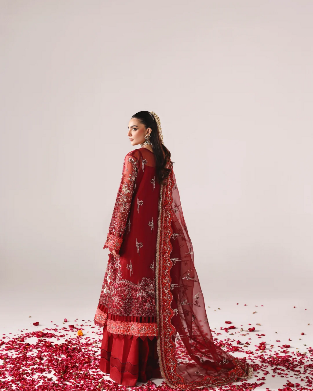 A woman wearing a traditional red embroidered outfit with a matching sheer dupatta stands in profile on a light studio background, with scattered red flower petals around her feet.