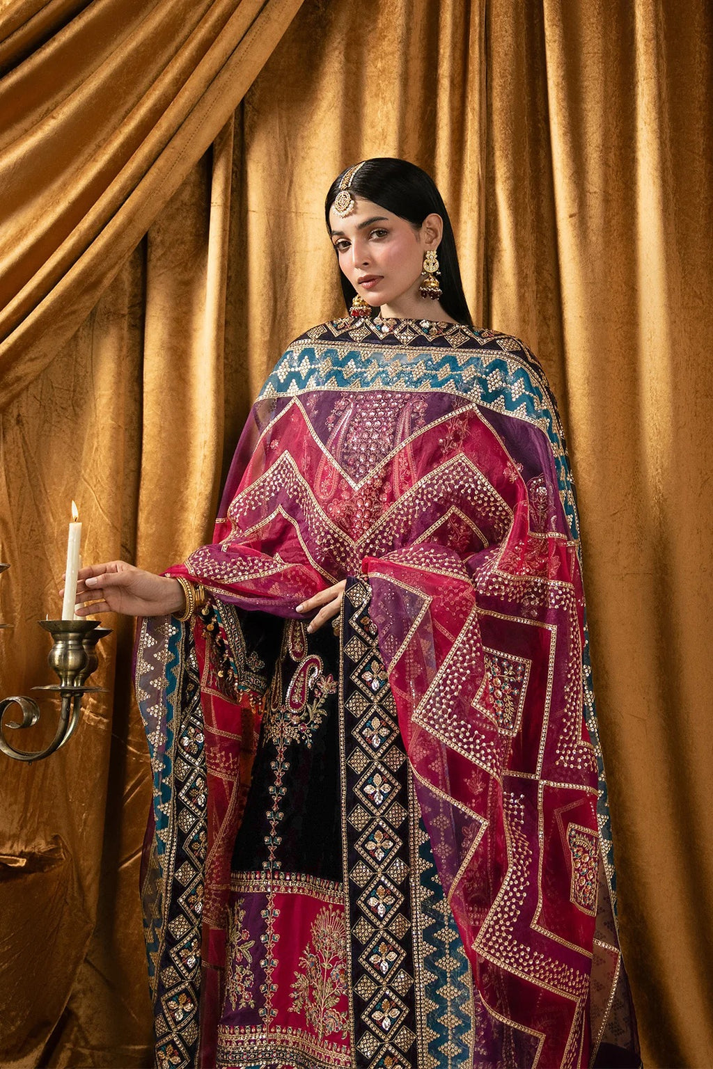 A woman models a traditional South Asian outfit featuring a black kurta heavily embellished with magenta, purple, and gold geometric patterns, paired with a matching sheer dupatta, set against a draped gold curtain background.