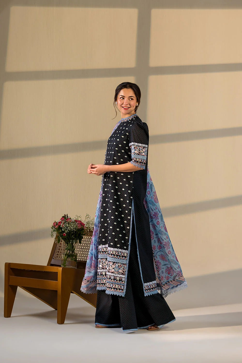 Woman in a black and blue traditional outfit standing in front of a beige wall with a wooden stool and flowers.