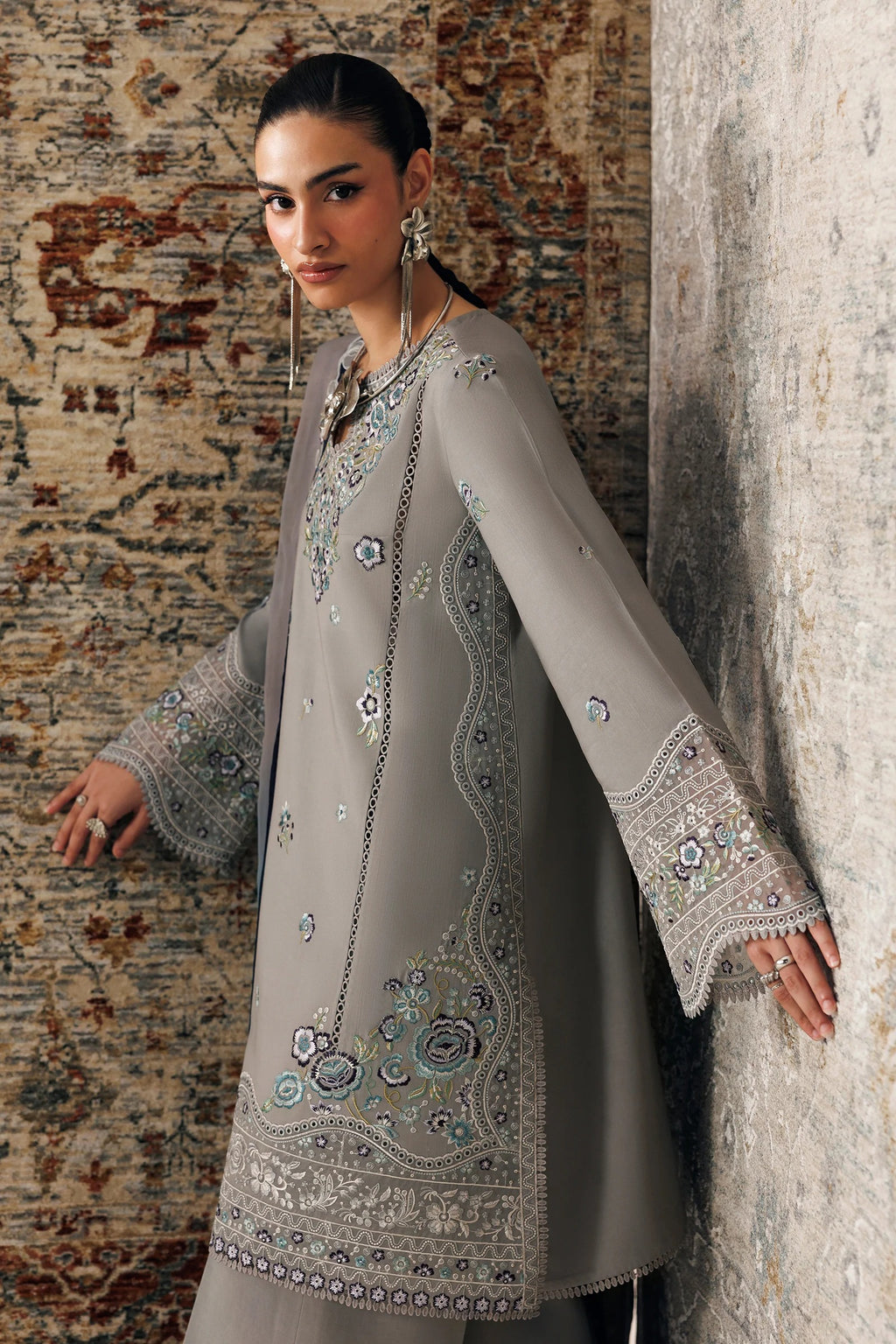 A woman models a light gray traditional long kurta with intricate silver and blue embroidery, standing against a textured wall next to a patterned rug.