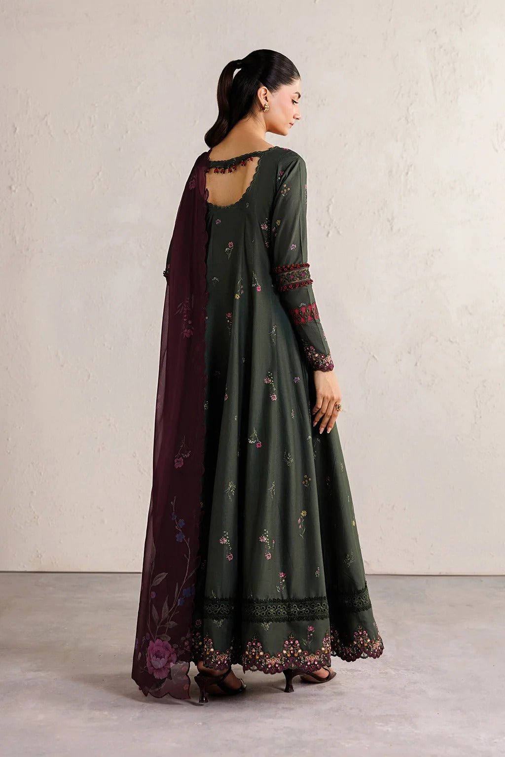 A woman modeling a dark olive green anarkali dress with floral embroidery, a deep neckline with lace trim, and a matching dark plum dupatta, shown from the back against a neutral textured wall.