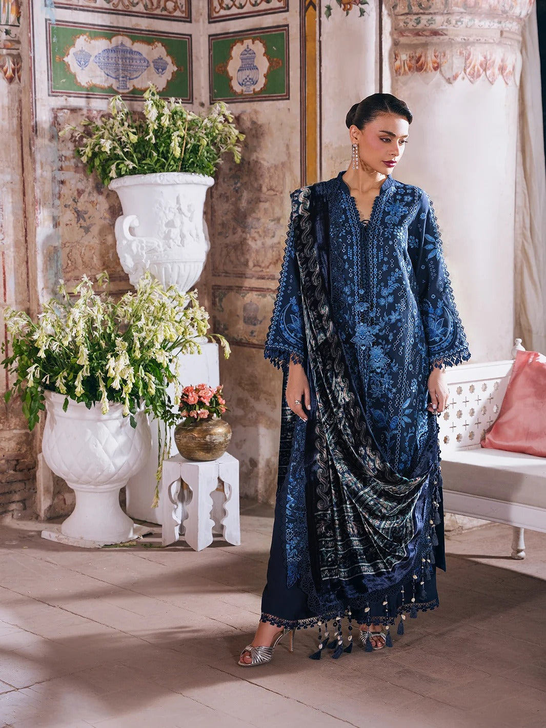 A woman models a traditional navy blue embroidered ethnic outfit with a matching dupatta in an ornate indoor setting with classical wall details, large white planters, and a bench.