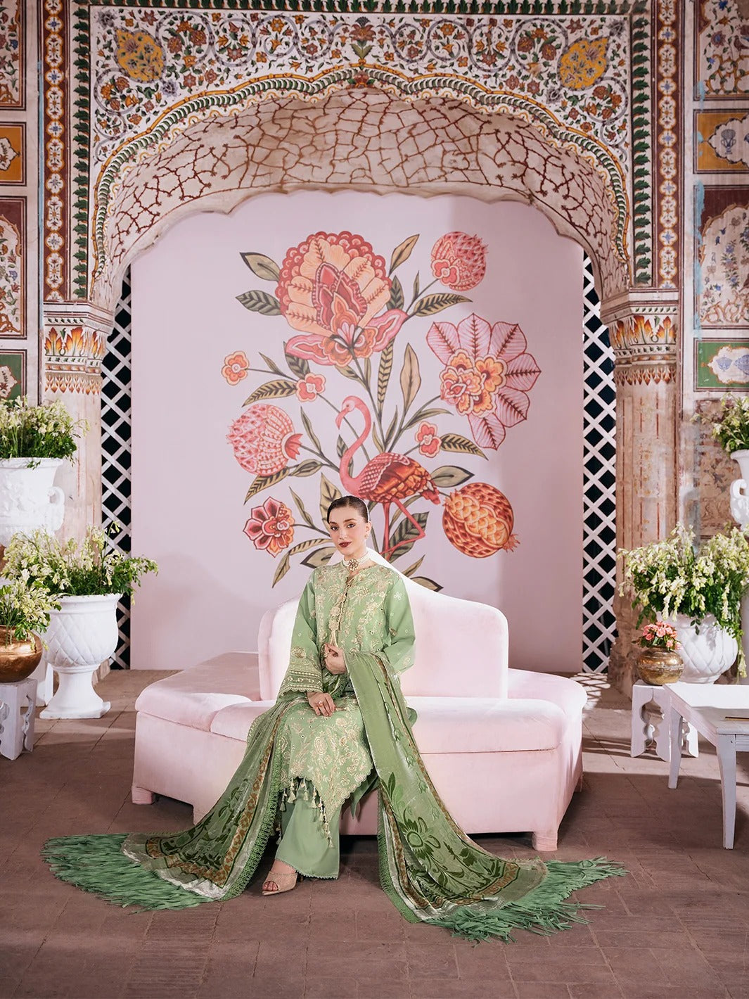 A woman wearing a green embroidered traditional outfit sits on a pale pink sofa in an ornate interior with a floral archway backdrop.
