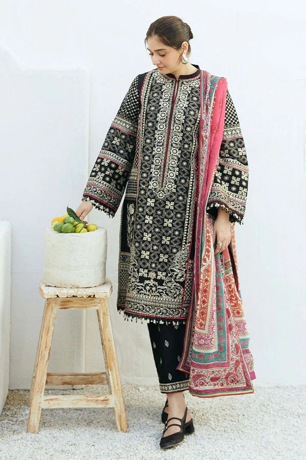 A woman models a traditional three-piece salwar kameez suit in black, featuring intricate white and red floral embroidery, paired with a pink and red patterned dupatta, standing next to a small wooden stool with a basket of lemons.