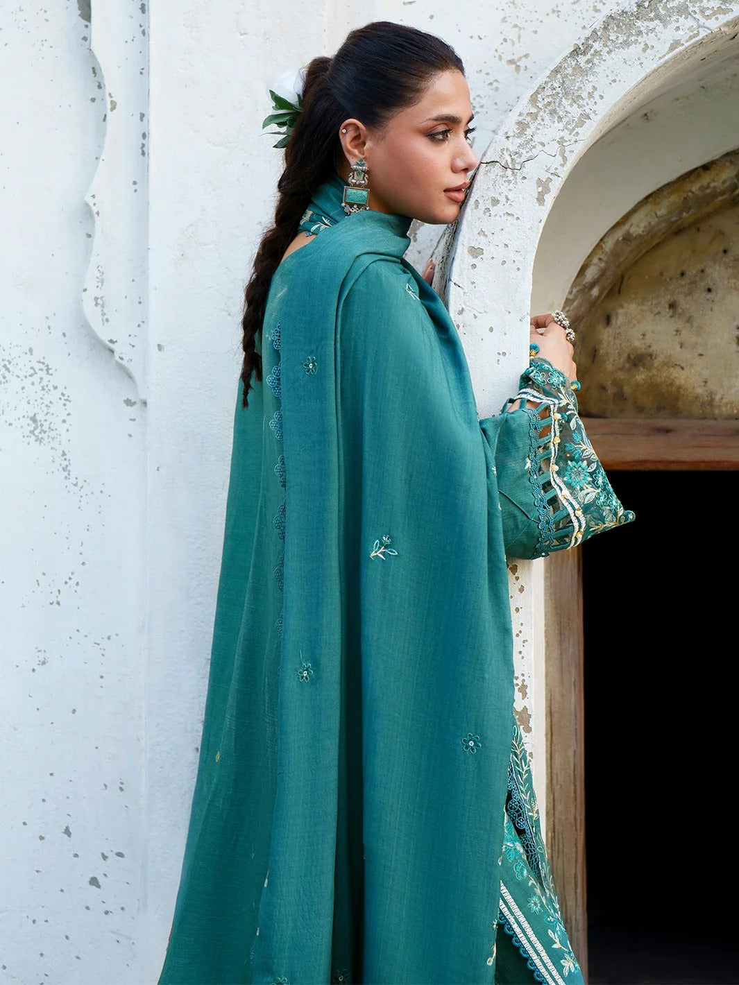 A woman wearing a teal green embroidered traditional outfit with a matching long cape, standing beside a rustic white stucco archway doorway.