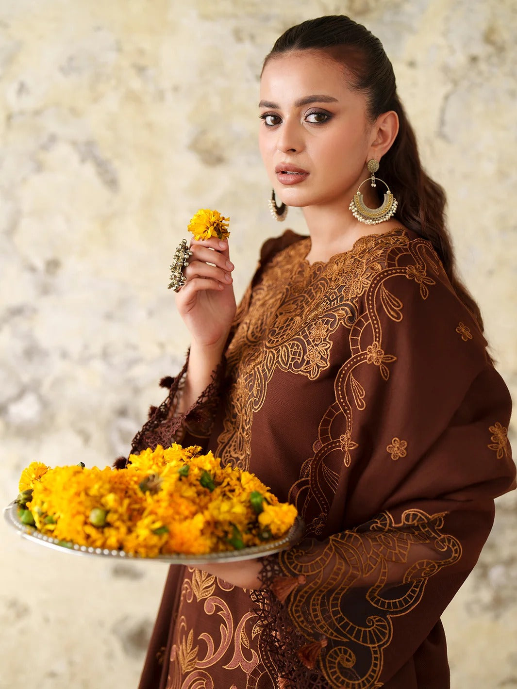A woman wearing a brown embroidered traditional outfit holds a silver tray filled with yellow marigold flowers, with one flower near her face, against a textured beige wall.
