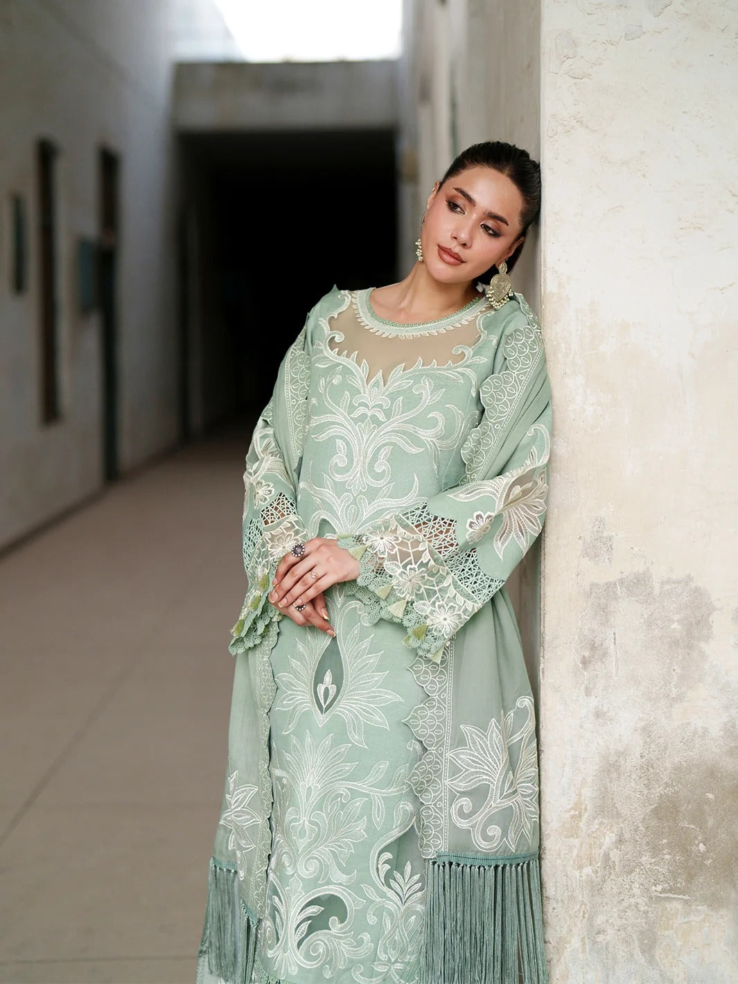 A woman wearing a mint green embroidered traditional outfit with white floral patterns and lace details, leaning against a textured wall in a narrow hallway.