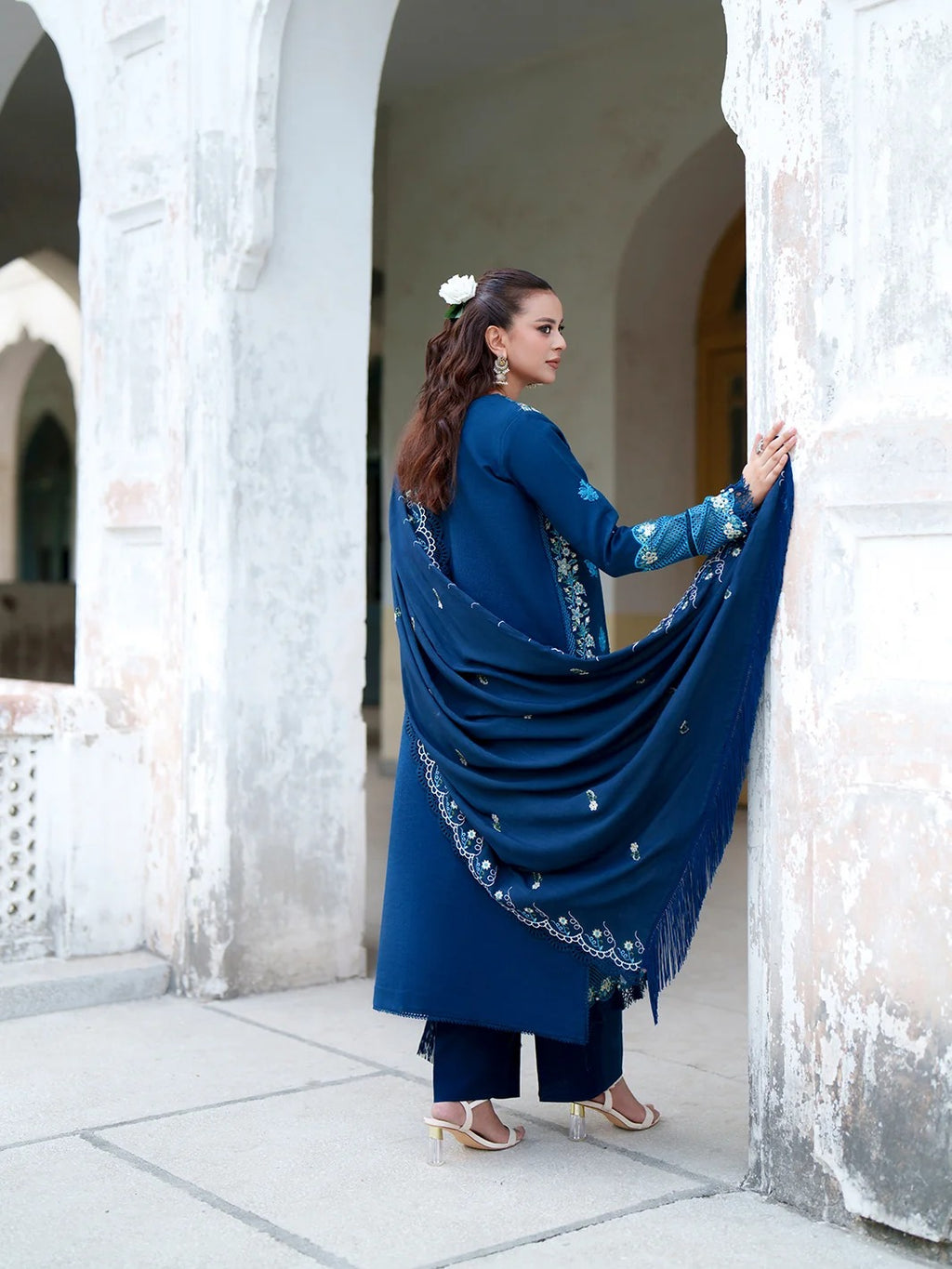 A woman wearing a deep blue traditional outfit with matching dupatta and dark blue pants, standing in a stone corridor with arches. She is holding out the dupatta to show its embroidered border.