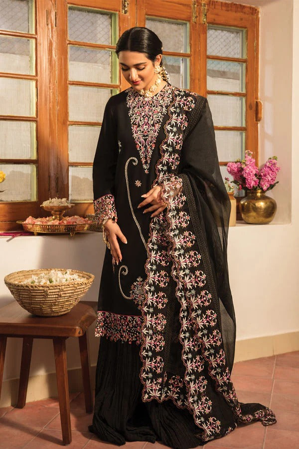 A woman models a black traditional South Asian outfit with elaborate pink and white floral embroidery, standing in a warm-toned room with wooden windows and a basket on a table.