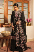 A woman models a black traditional South Asian outfit with elaborate pink and white floral embroidery, standing in a warm-toned room with wooden windows and a basket on a table.