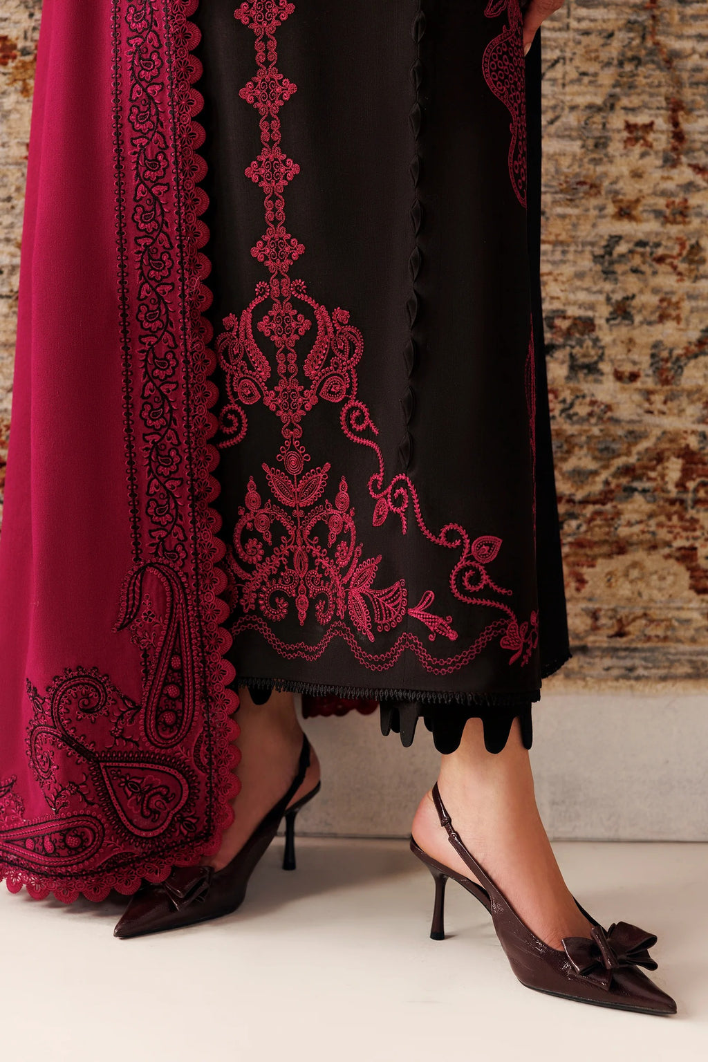 A close-up of a person wearing a black traditional outfit with ornate magenta embroidery and a matching sheer dupatta, paired with dark slingback heels, standing against a textured stone wall.