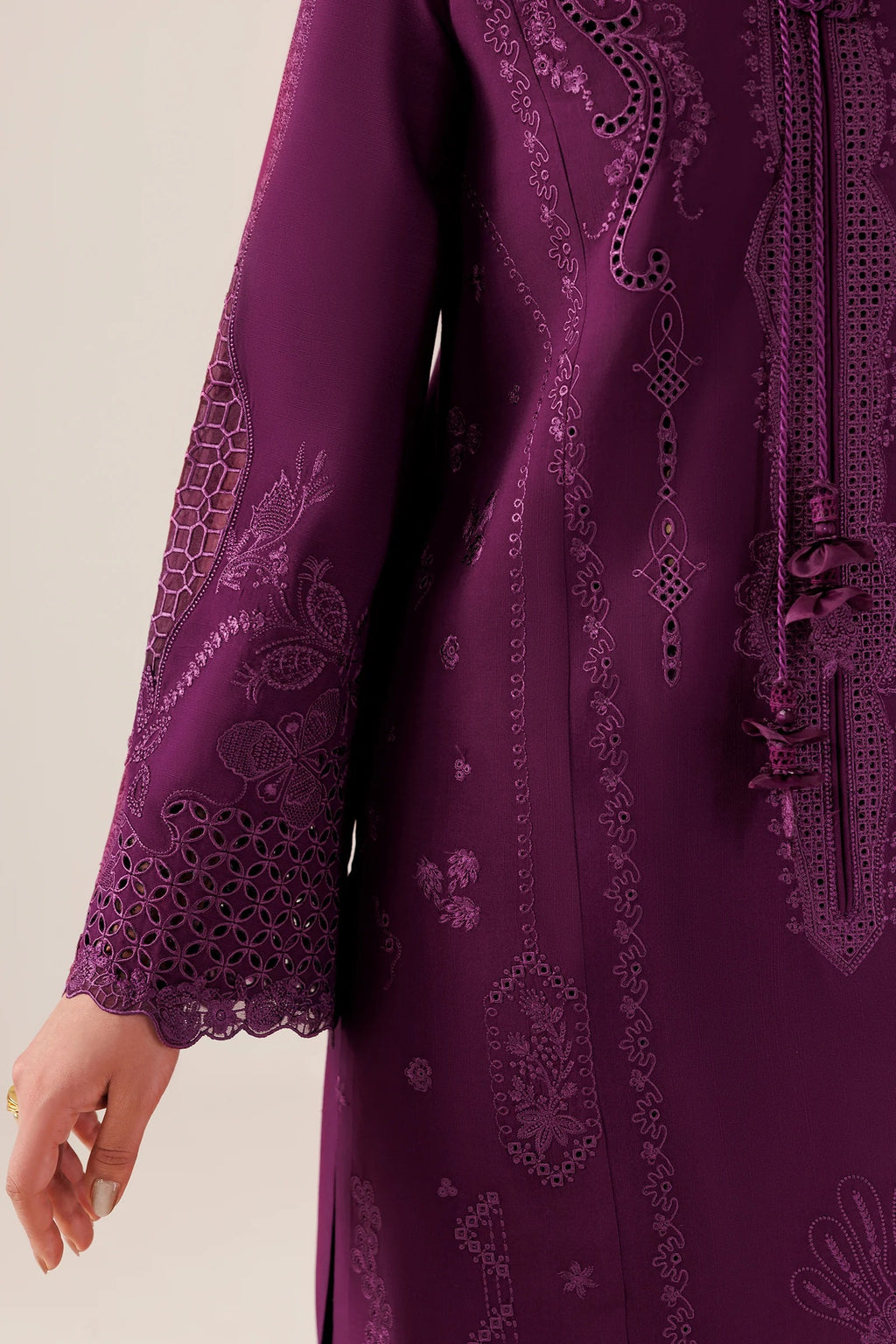 A close-up of a deep purple embroidered tunic or kurta with intricate floral and paisley embroidery and lace detailing on the sleeve and front panel, shown against a plain light beige background.