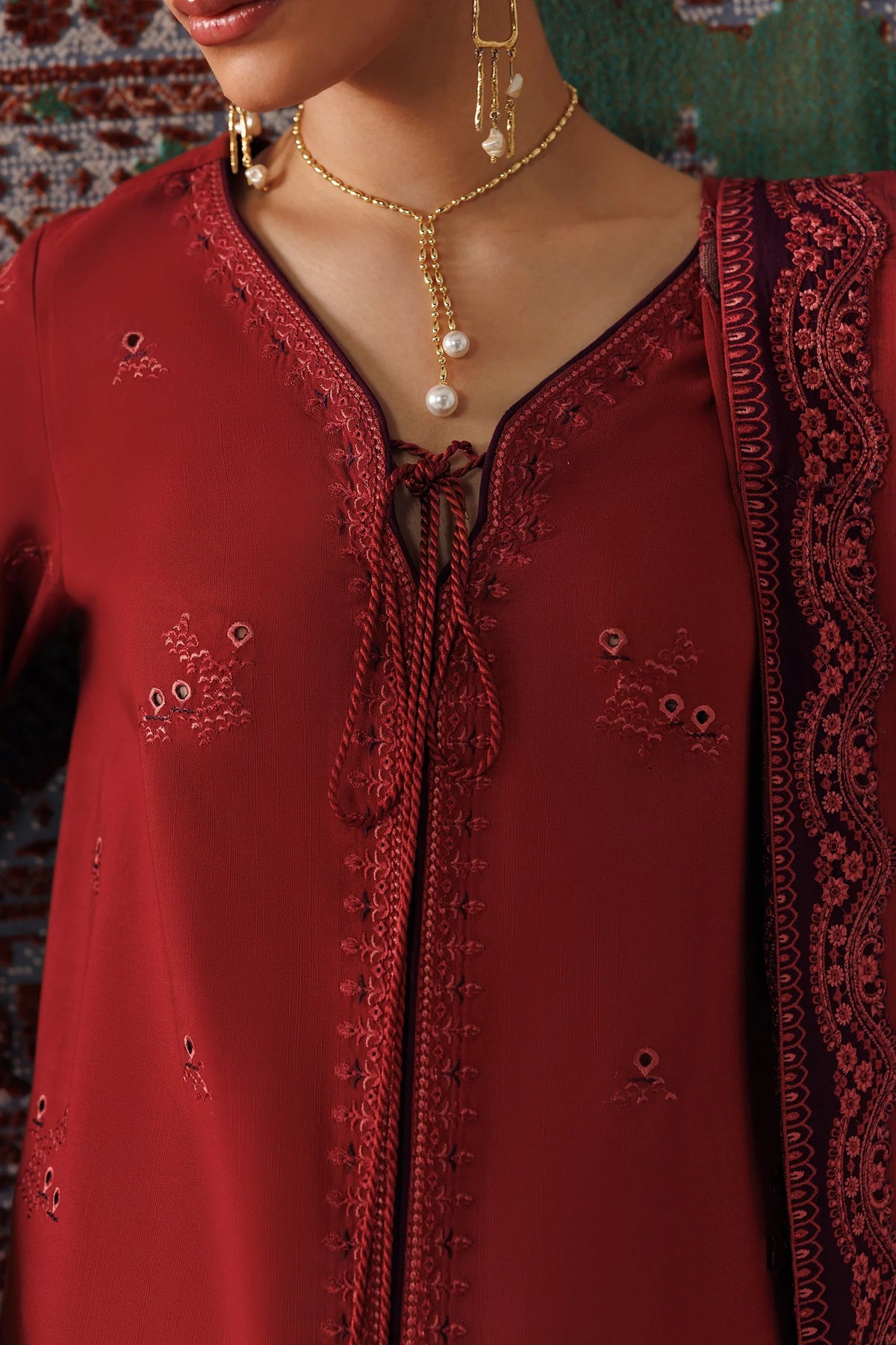 A close-up of a woman wearing a deep red embroidered traditional outfit with a V-neckline, tie-front detail, and matching embroidered dupatta. Gold jewelry is also visible.