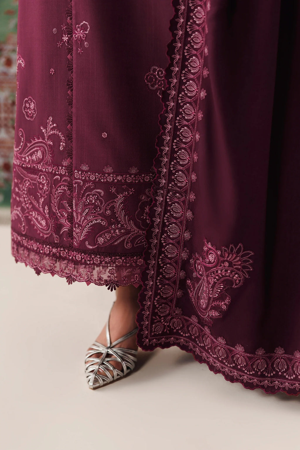 A close-up of a deep burgundy maxi dress with intricate floral embroidery and lace borders, paired with metallic silver pointed-toe flats.
