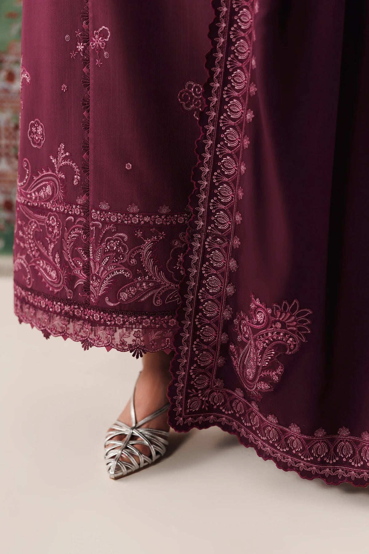 A close-up of a deep burgundy maxi dress with intricate floral embroidery and lace borders, paired with metallic silver pointed-toe flats.