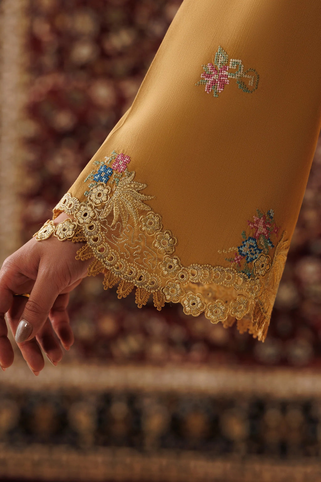 A close-up of a mustard yellow embroidered fabric sleeve or garment hem, held by a hand. The sleeve features delicate floral embroidery and a scalloped lace cuff, set against a blurred patterned rug background.