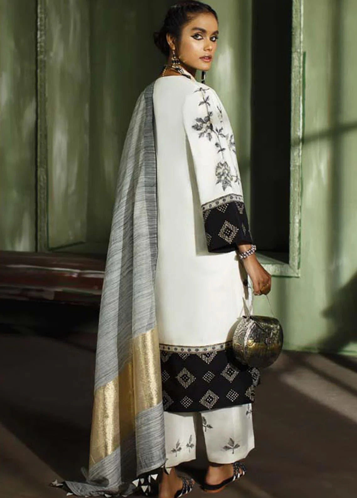 A woman models a traditional South Asian outfit featuring a white kurta with black floral embroidery and a black patterned border, paired with a white dupatta that has a gray and gold striped design. She holds a metallic gold handbag in a dimly lit, moody interior setting.