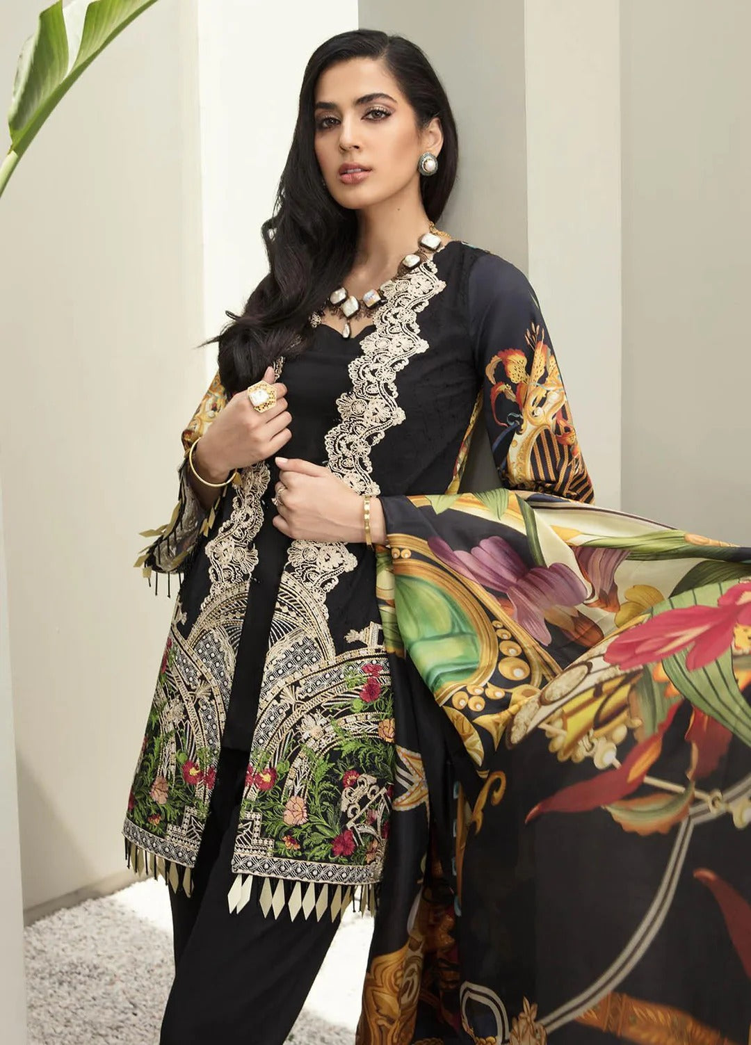A woman models a black three-piece traditional outfit with intricate white floral embroidery and a matching printed dupatta featuring colorful botanical and gold ornamental motifs.