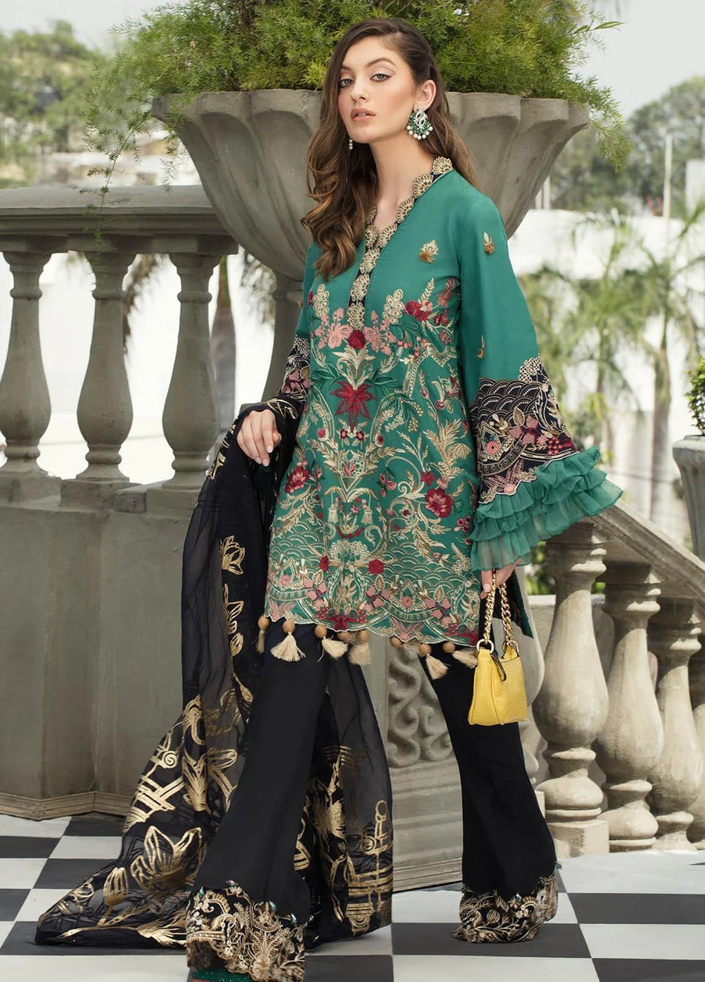 A woman models a traditional three-piece salwar kameez suit. The outfit features a teal green kameez top with intricate floral embroidery, paired with wide-leg black flared pants and a matching black dupatta decorated with gold and red patterns.