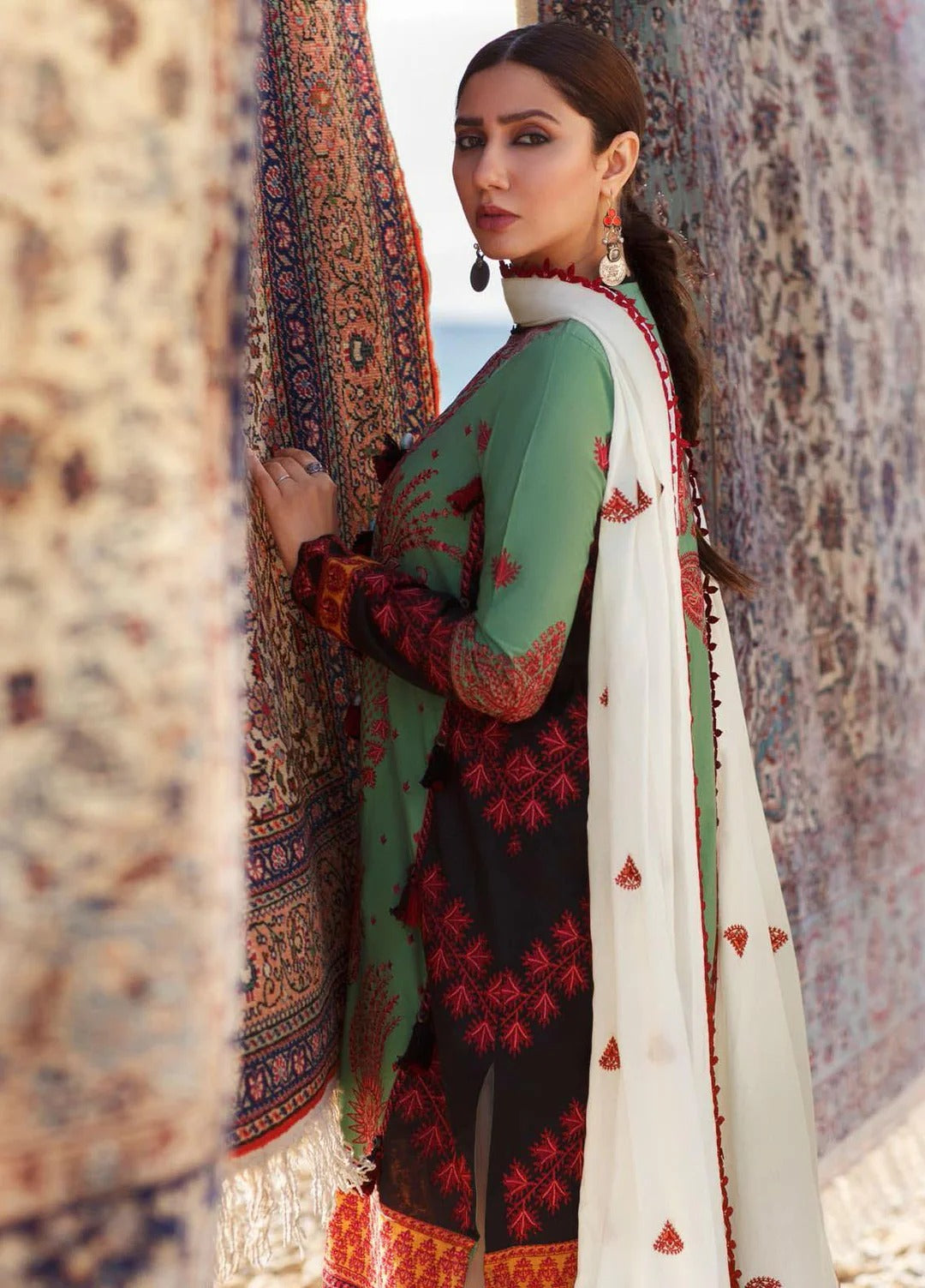 A woman models a traditional South Asian salwar suit set featuring a sage green top with red floral embroidery, a black bottom with red embroidery, and a white dupatta with red accents, framed by patterned rugs.