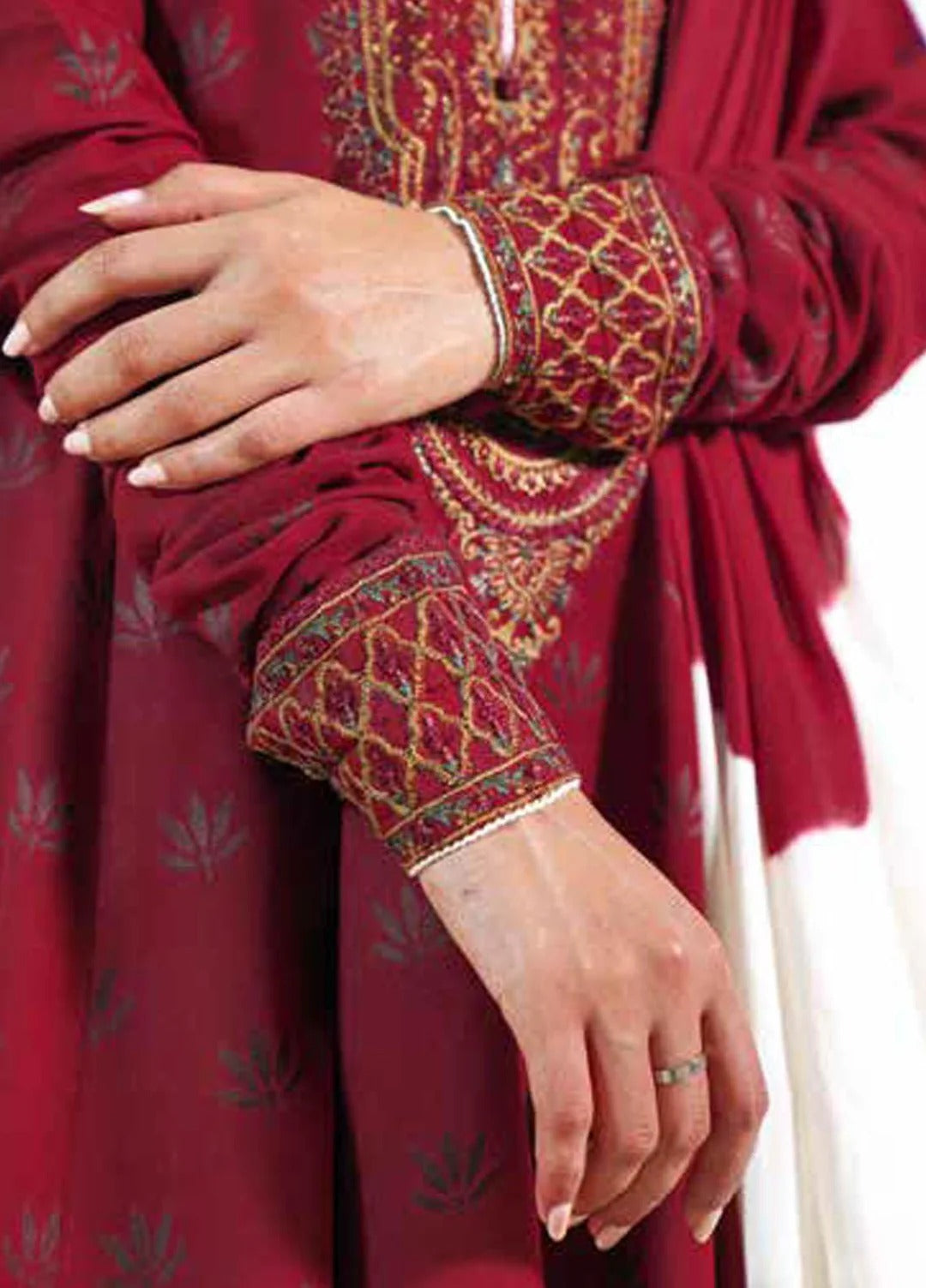 A close-up of a person wearing a deep red traditional outfit with ornate gold and green embroidery on the sleeves and neckline, along with a matching draped scarf.