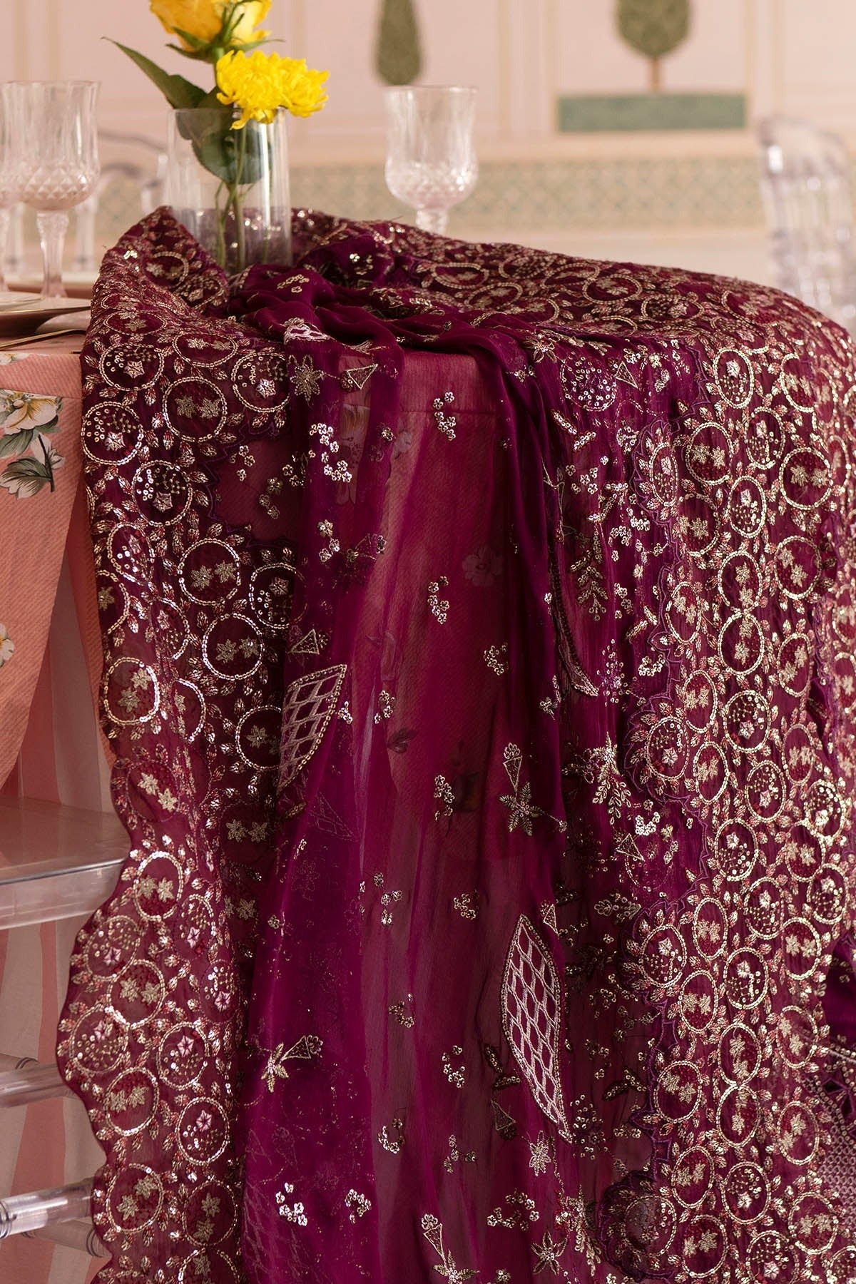 A deep magenta purple embroidered fabric, likely a dupatta or scarf, draped over a chair in an indoor setting. The sheer mesh fabric features intricate metallic gold and silver floral and geometric patterns.
