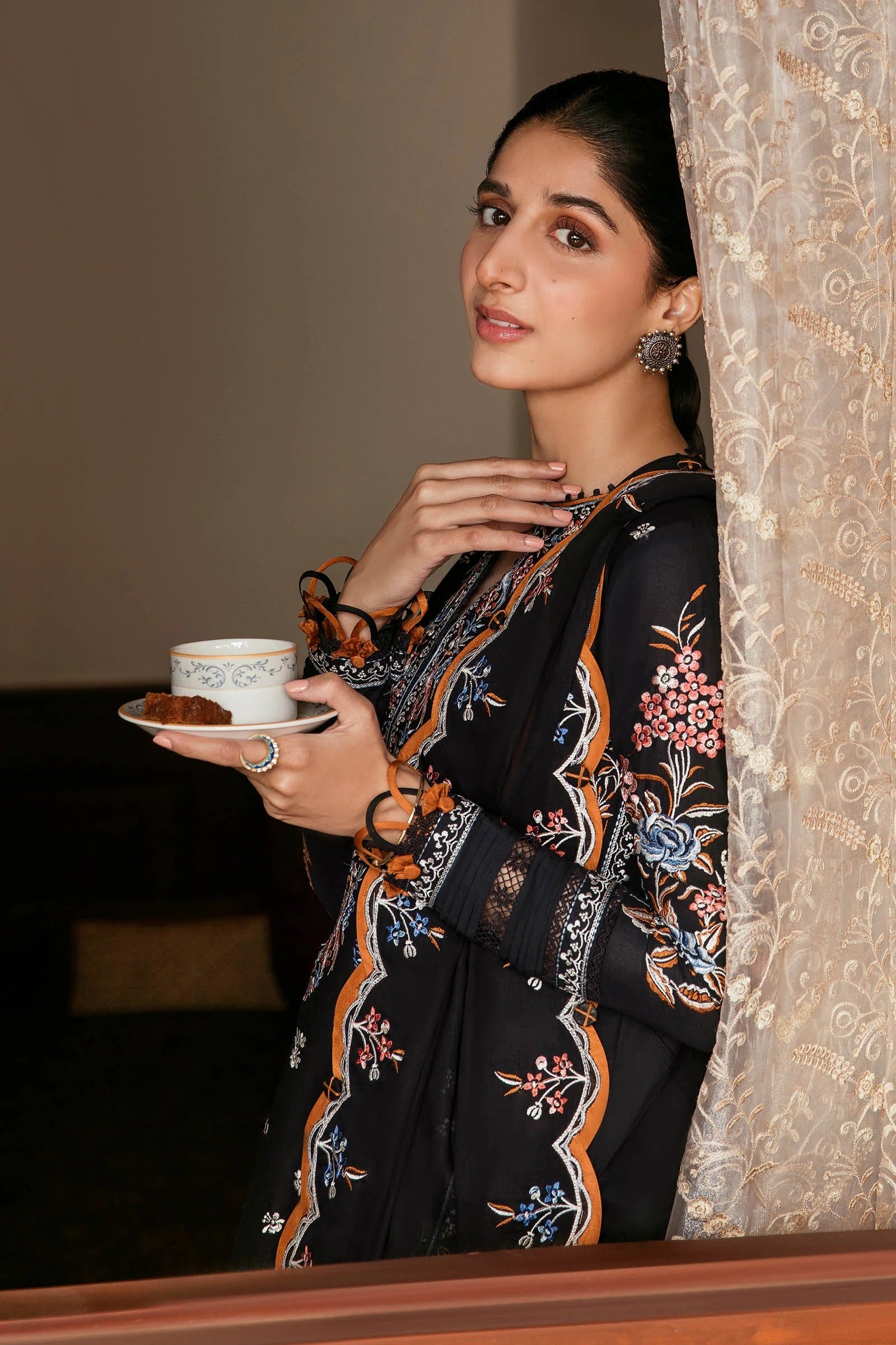 Woman holding a cup and saucer, standing behind a lace curtain.