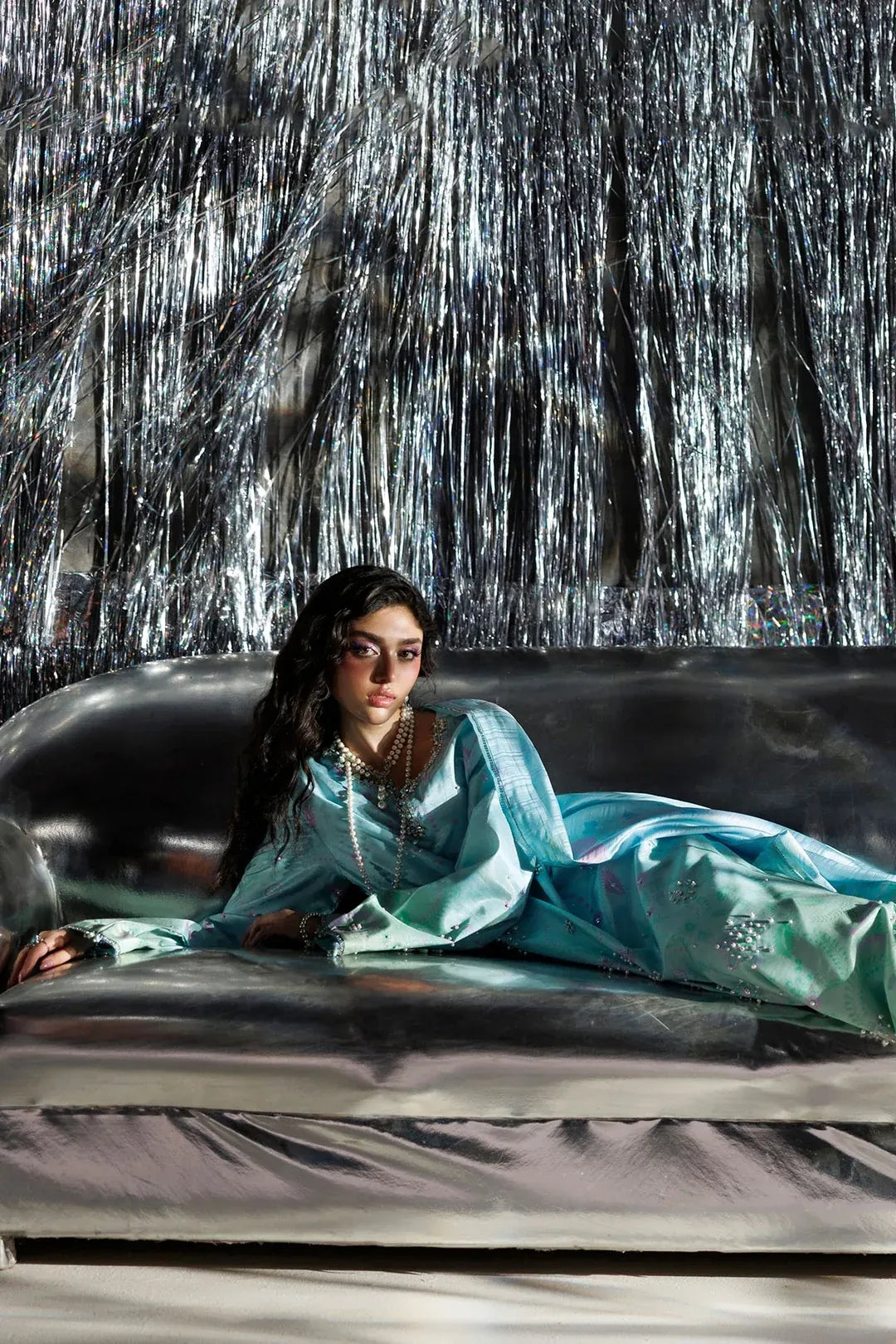 A woman reclines on a metallic sofa wearing a flowing light blue traditional outfit and layered necklaces, posed against a reflective silver tinsel backdrop.