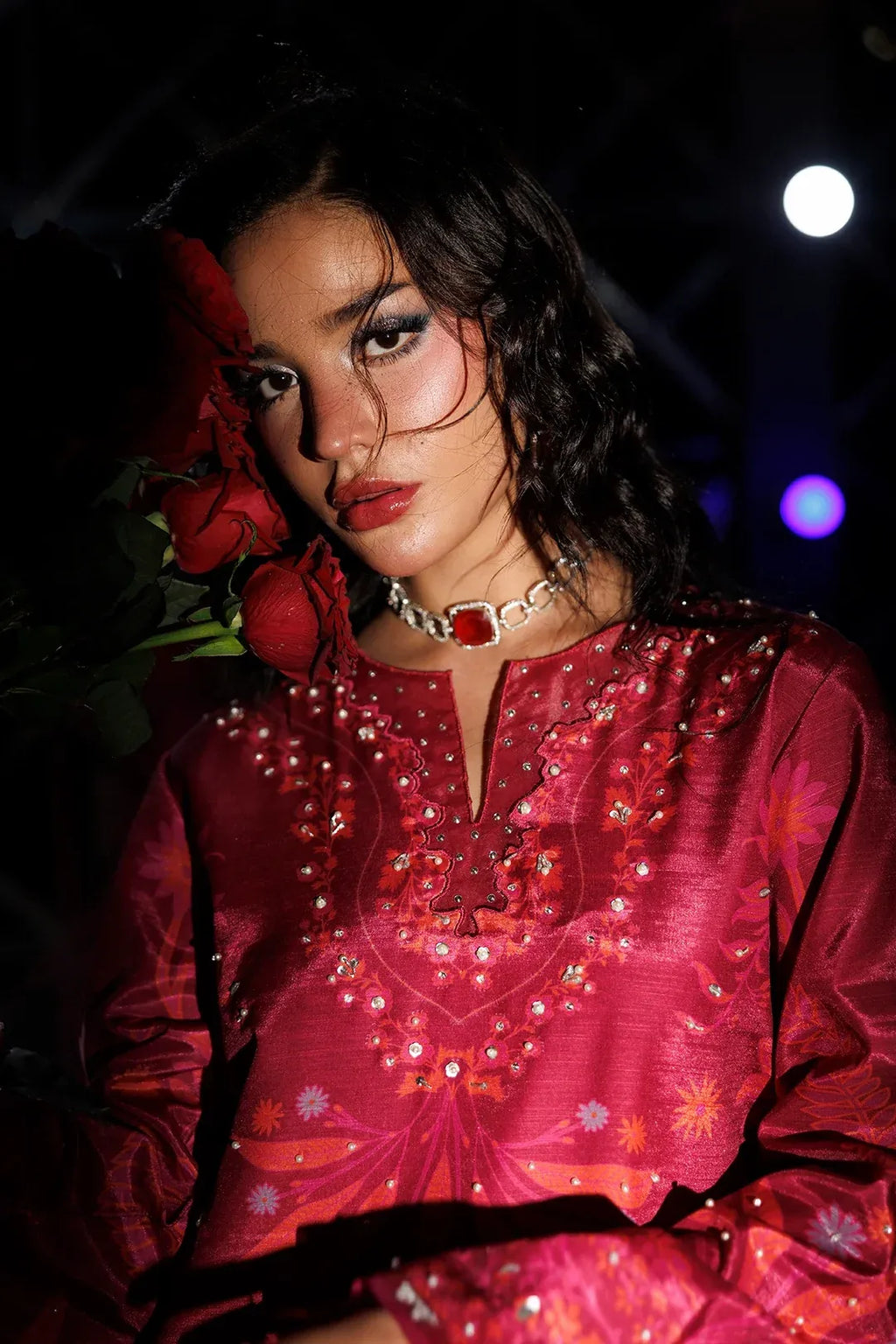 A woman wearing a deep red embellished kurta with floral embroidery and beadwork, styled with a red rose in her hair and a statement necklace, set against a dark background with stage-like lights.