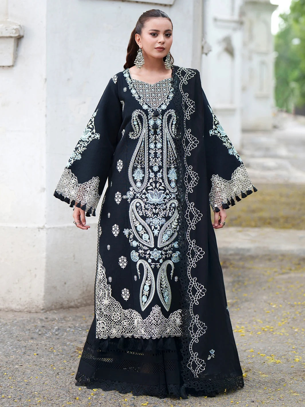 A woman models a long black embroidered traditional dress with intricate silver and white paisley and floral patterns, standing outdoors against a light-colored stone wall.