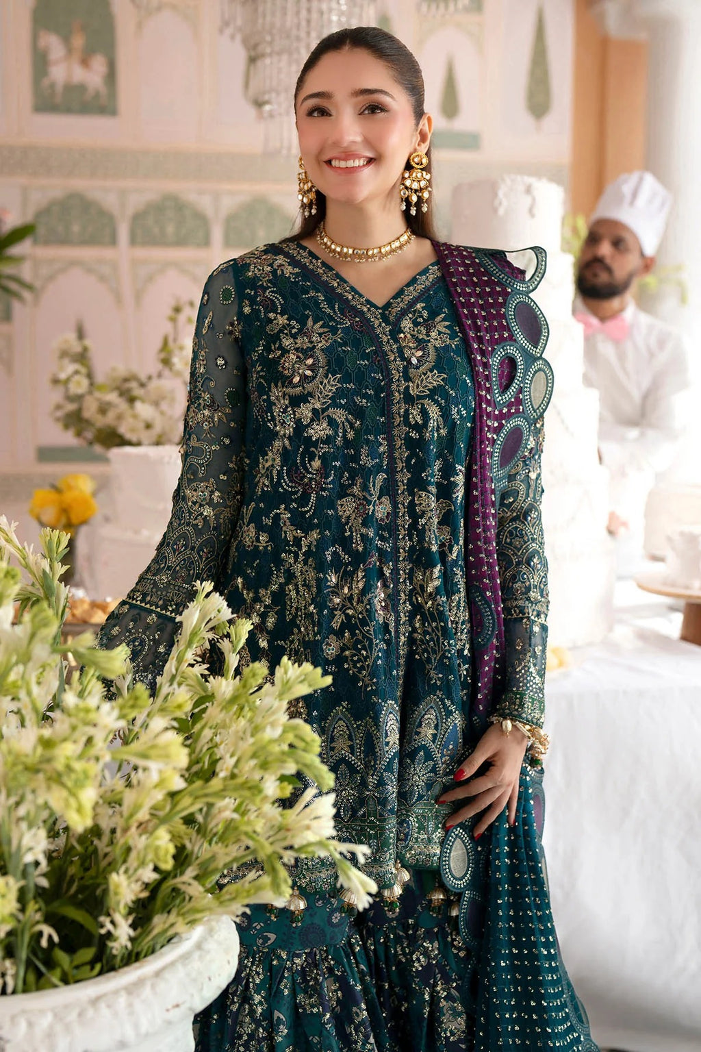 A woman models a dark teal green heavily embroidered traditional outfit with gold detailing, a purple patterned dupatta, and gold jewelry in an indoor event setting.