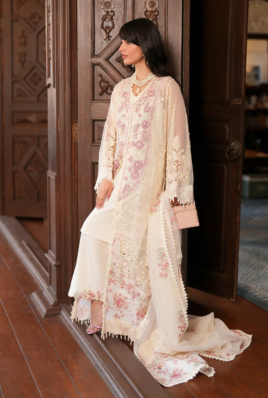 A woman models a traditional three-piece salwar kameez suit in cream with pink floral embroidery, standing in front of a dark wooden door.