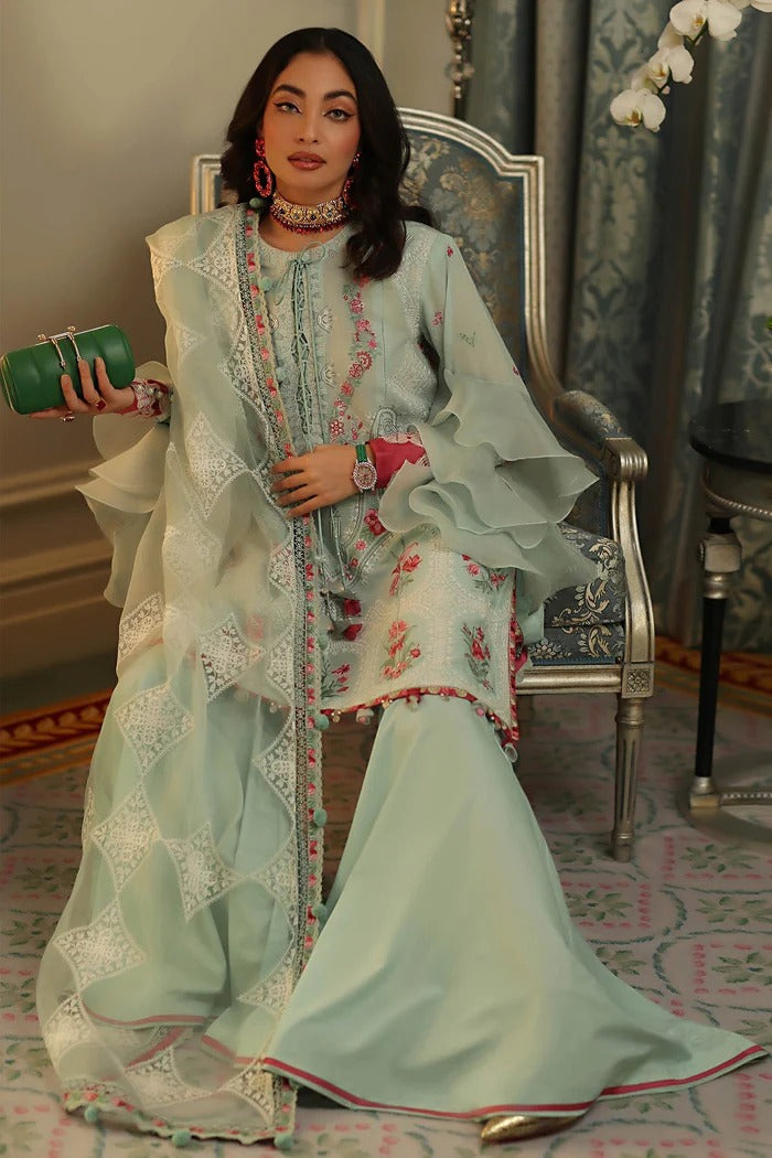 A woman seated in an ornate chair wearing a light mint green traditional outfit with white embroidered lace panels and pink floral embroidery. She holds a green clutch in a decorated indoor setting with patterned carpet and curtains.