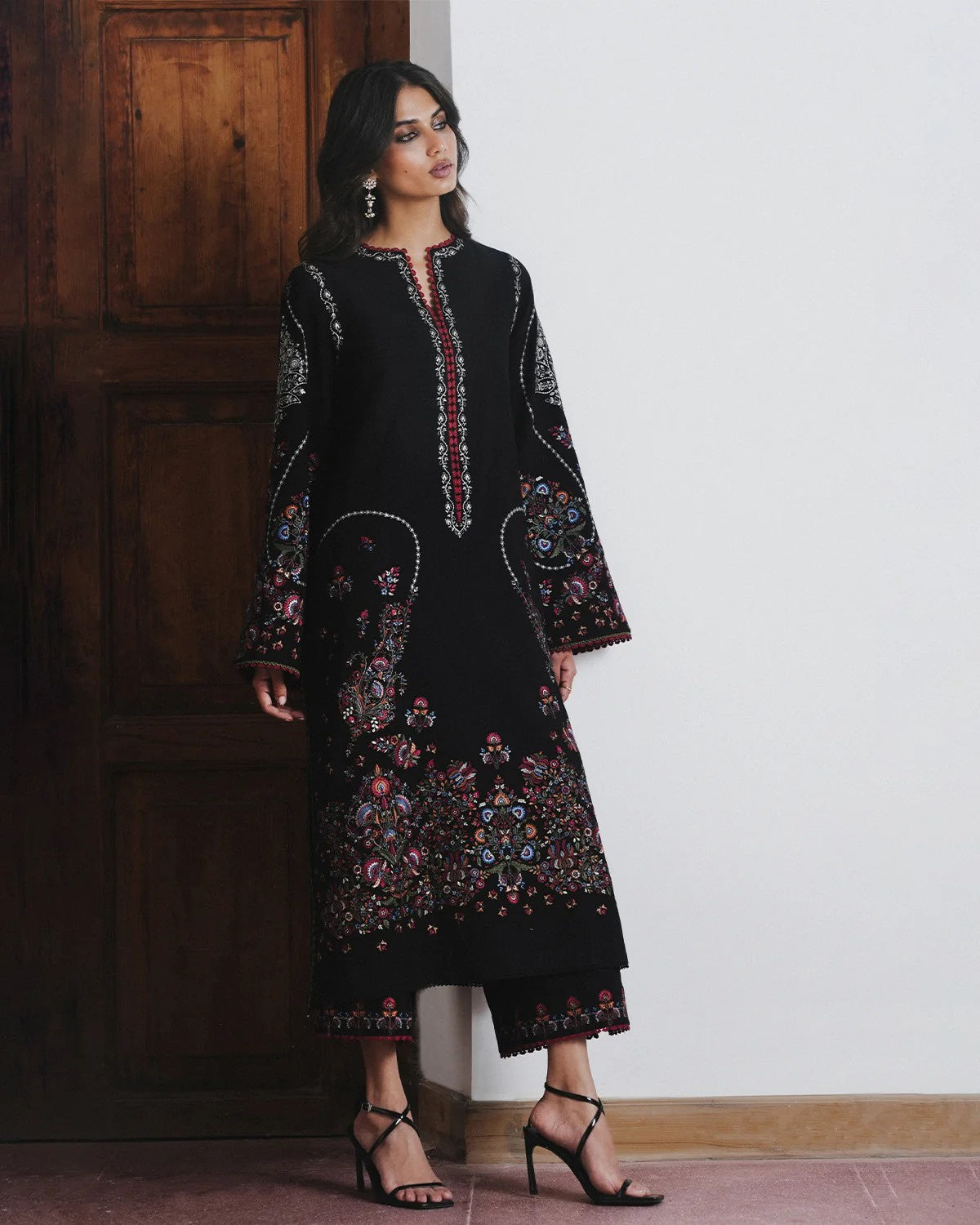 A woman models a black long kurta with colorful floral embroidery and beadwork details, paired with matching black pants and black high-heeled sandals, standing in front of a wooden door and a white wall.