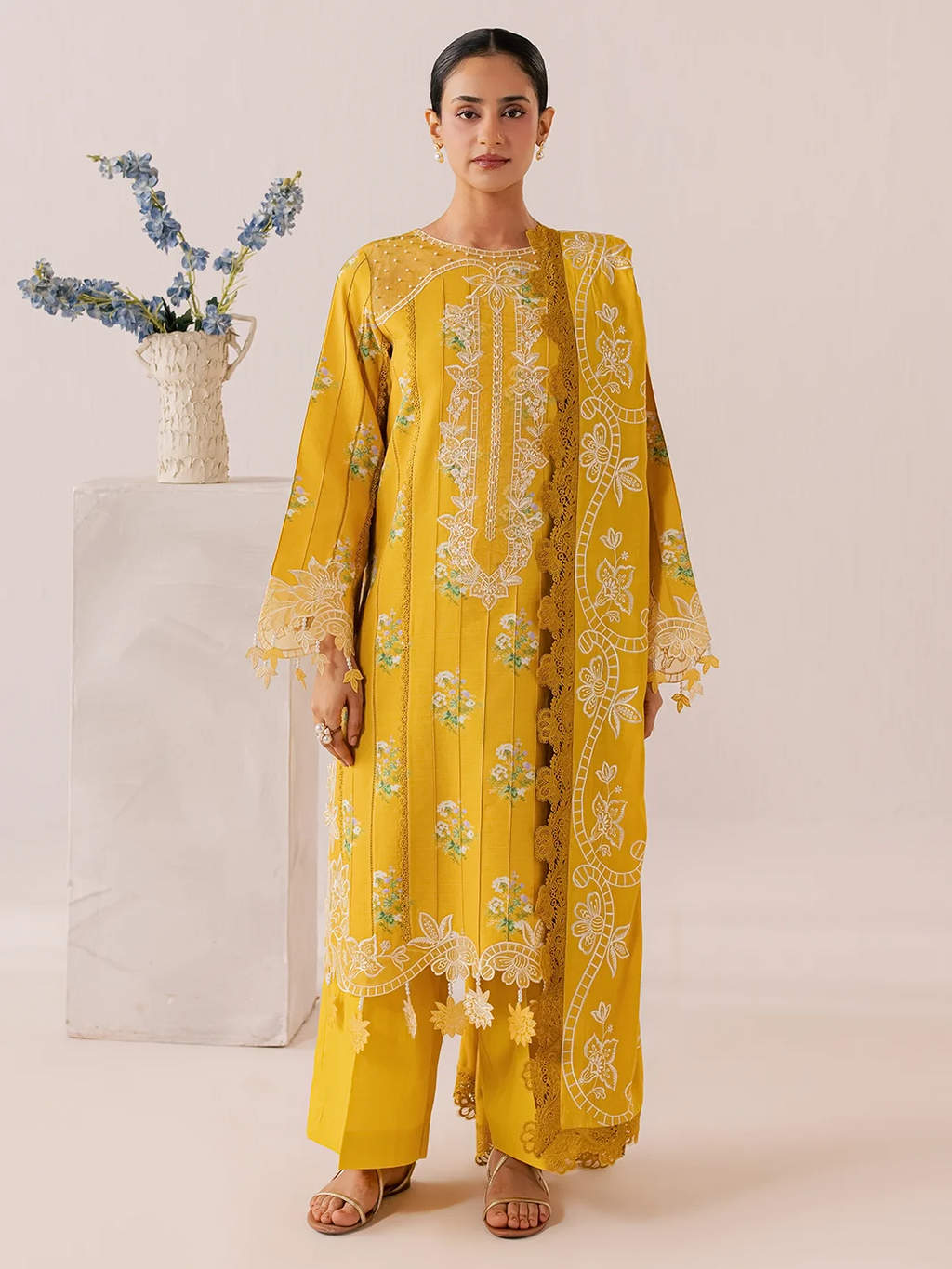 A woman models a bright yellow traditional outfit with intricate white and green floral embroidery, standing in front of a plain beige background next to a white pedestal with blue flowers.