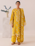A woman models a bright yellow traditional outfit with intricate white and green floral embroidery, standing in front of a plain beige background next to a white pedestal with blue flowers.