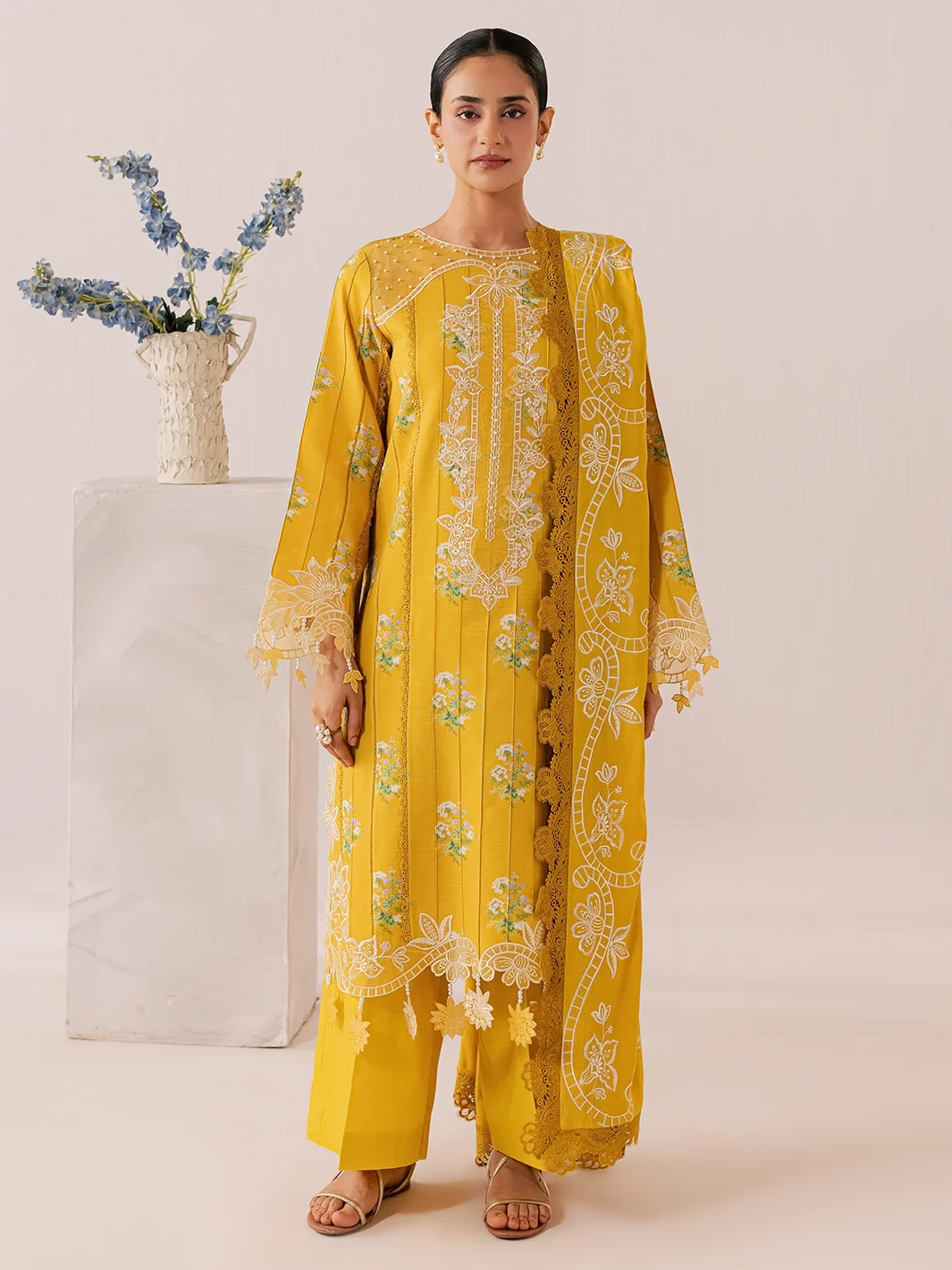 A woman models a bright yellow traditional outfit with intricate white and green floral embroidery, standing in front of a plain beige background next to a white pedestal with blue flowers.