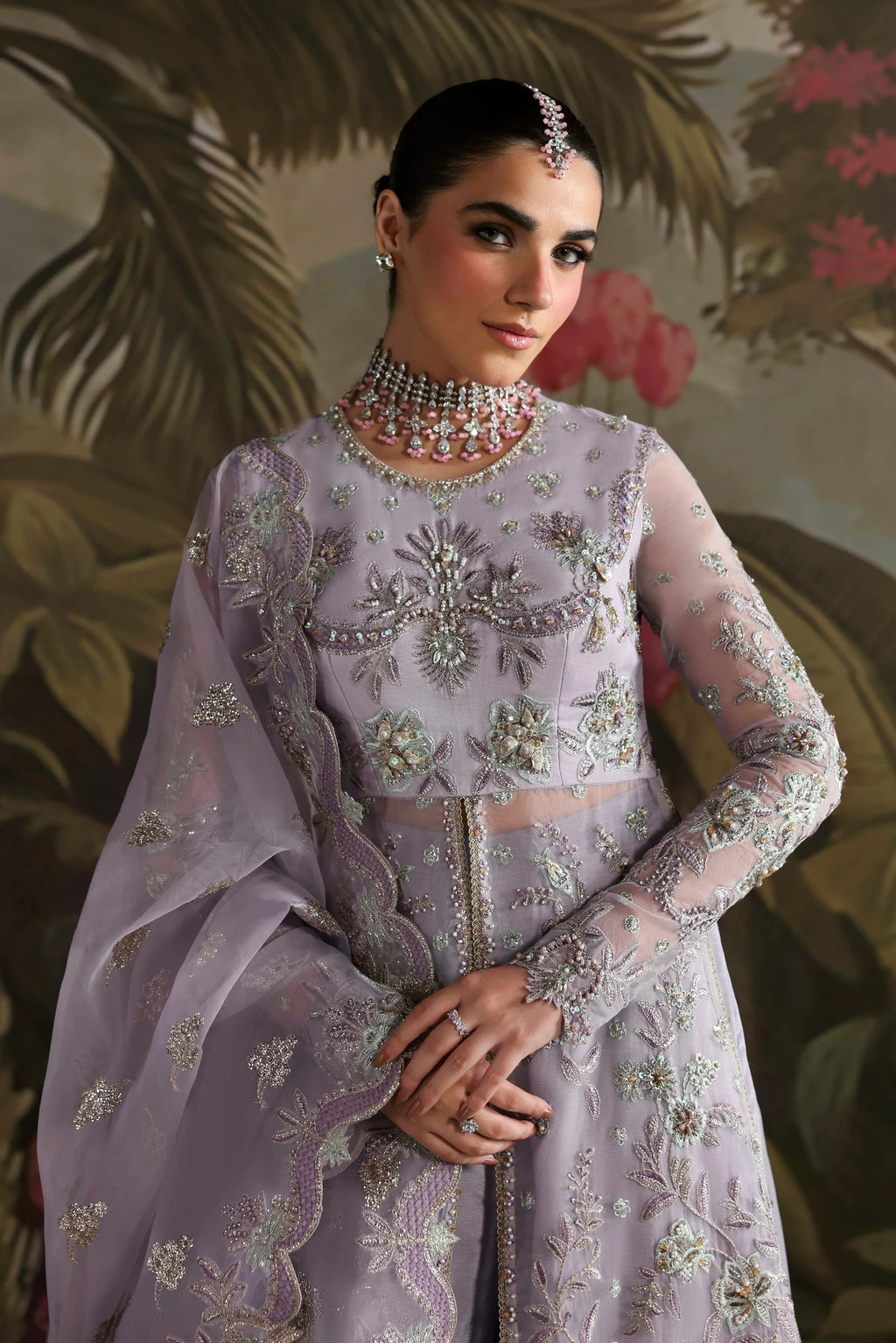 A woman modeling a traditional Indian outfit featuring a lavender-purple embroidered kurta with matching sheer dupatta and a heavily embellished silver and gold necklace, set against a dark tropical foliage background.