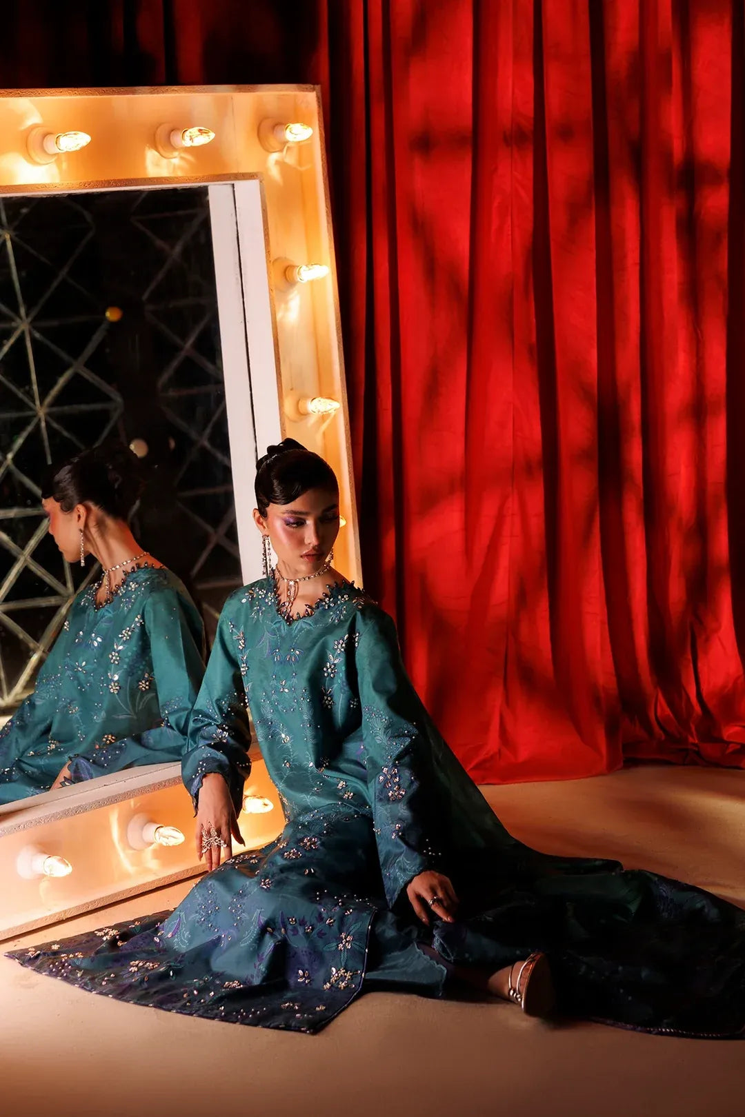 A woman wearing a teal embroidered traditional outfit sits on the floor in front of a red curtain and a lit mirror, with her reflection visible in the mirror.
