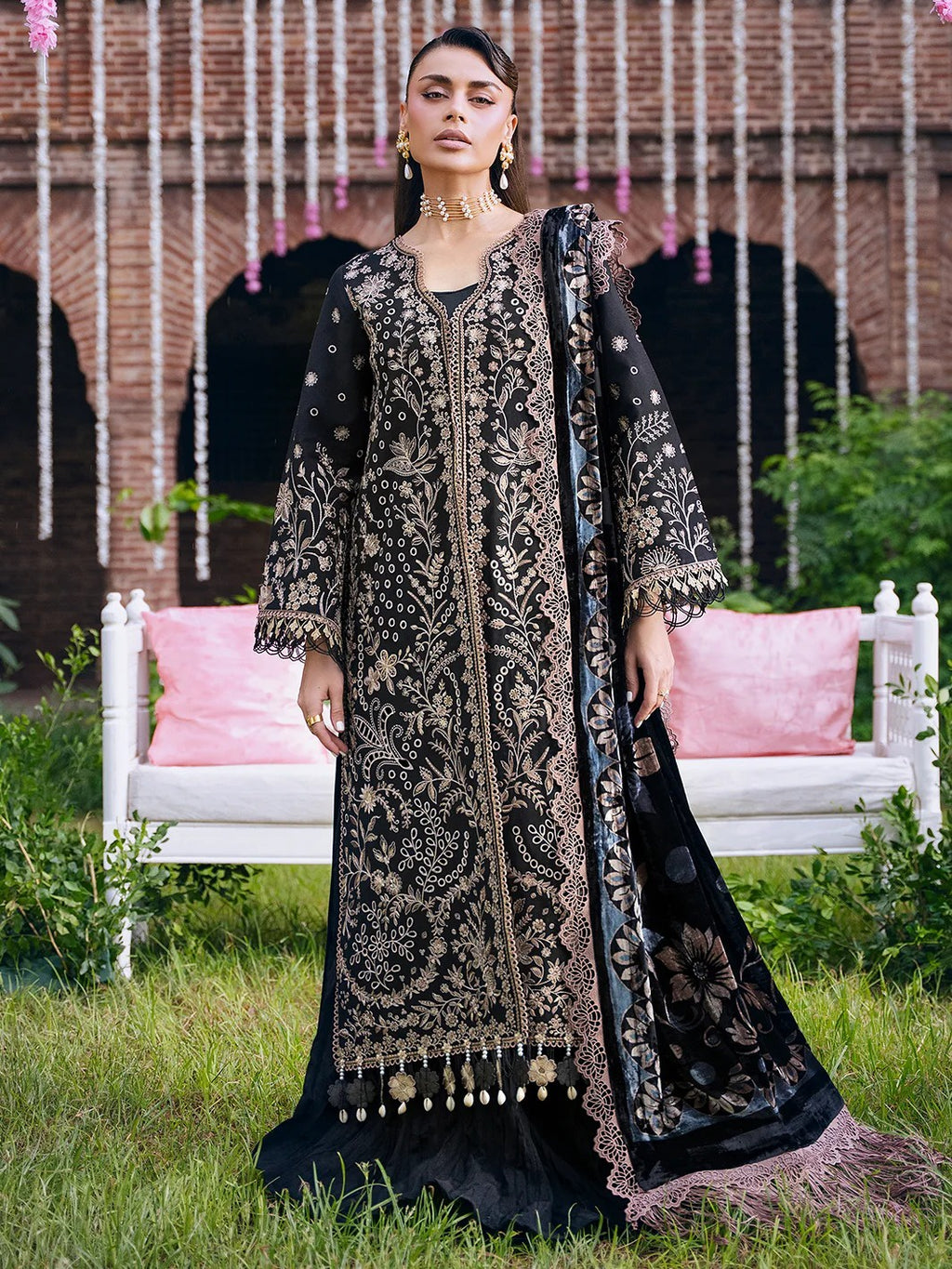 A woman models a black embroidered traditional outfit with gold detailing, featuring a long kurta, matching dupatta, and embellished tassel hem, set against a garden backdrop with a brick wall and white bench.