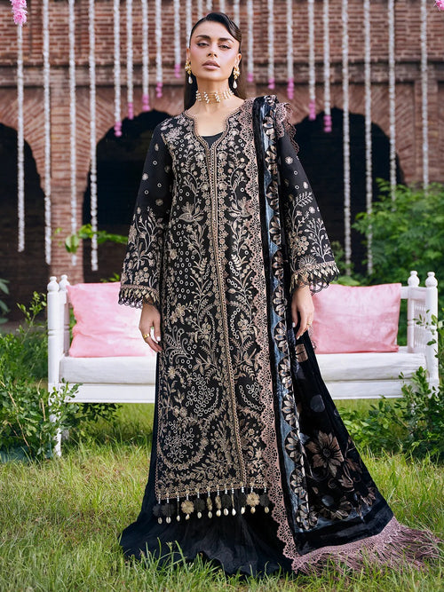 A woman models a black embroidered traditional outfit with gold detailing, featuring a long kurta, matching dupatta, and embellished tassel hem, set against a garden backdrop with a brick wall and white bench.