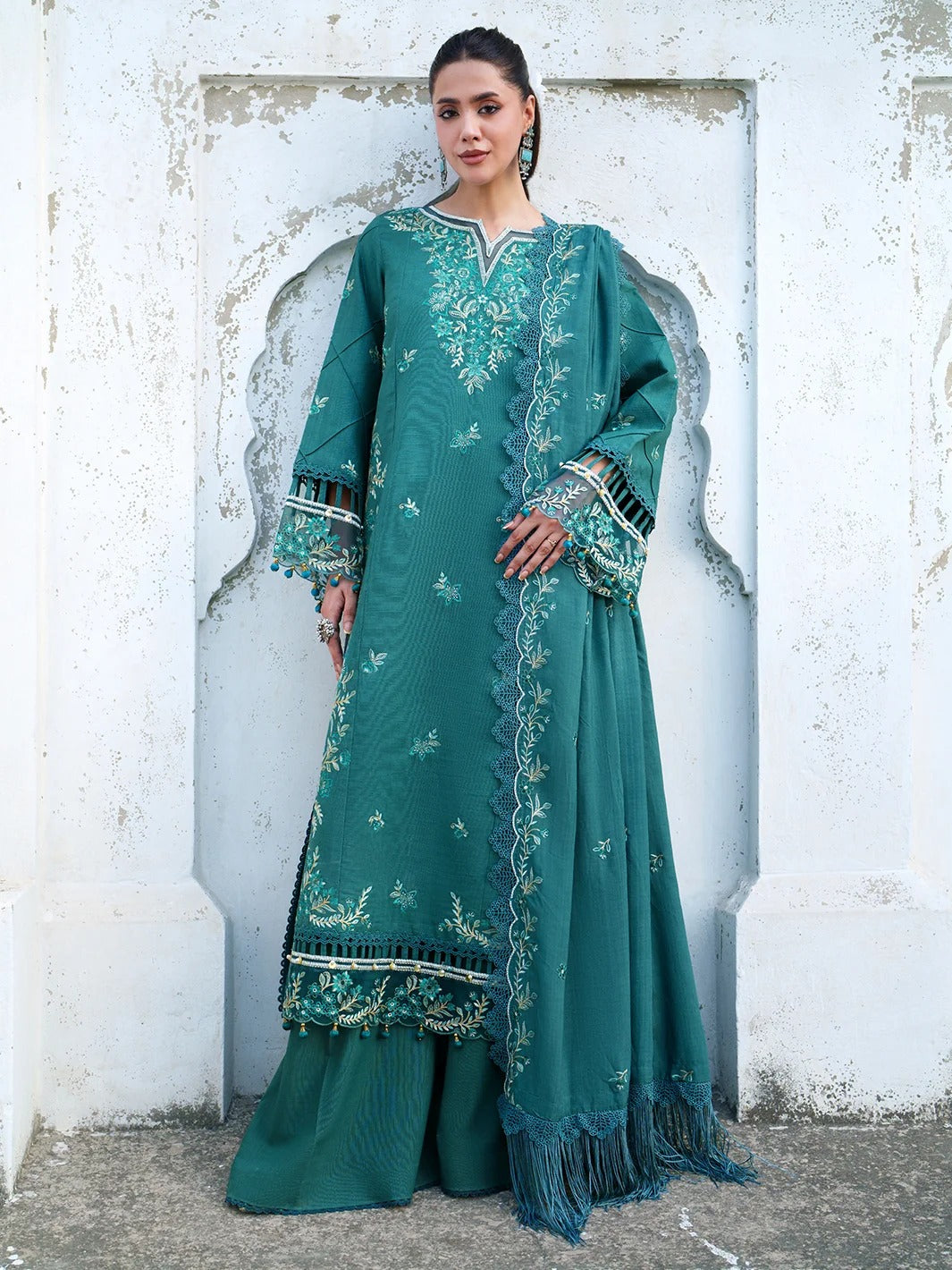 A woman models a traditional teal green embroidered kurta set with matching wide-leg pants and a fringed dupatta, standing against a white textured wall with a recessed arch design.