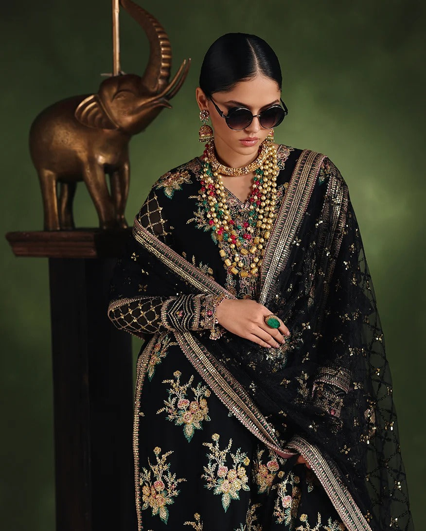 A woman models a black embroidered traditional outfit with a matching sheer dupatta, gold-toned jewelry, and dark sunglasses, posed in front of a dark green backdrop with a bronze elephant sculpture.